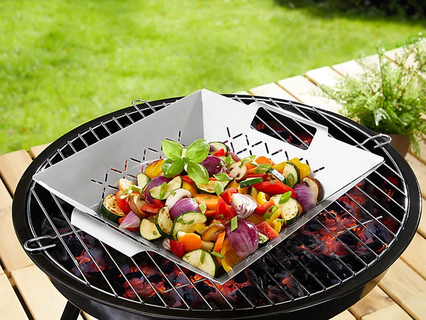 Gegrilltes Gemüse mit Basilikum in einem Grillkorb auf einem Grill, umgeben von üppigem Grün im Freien.