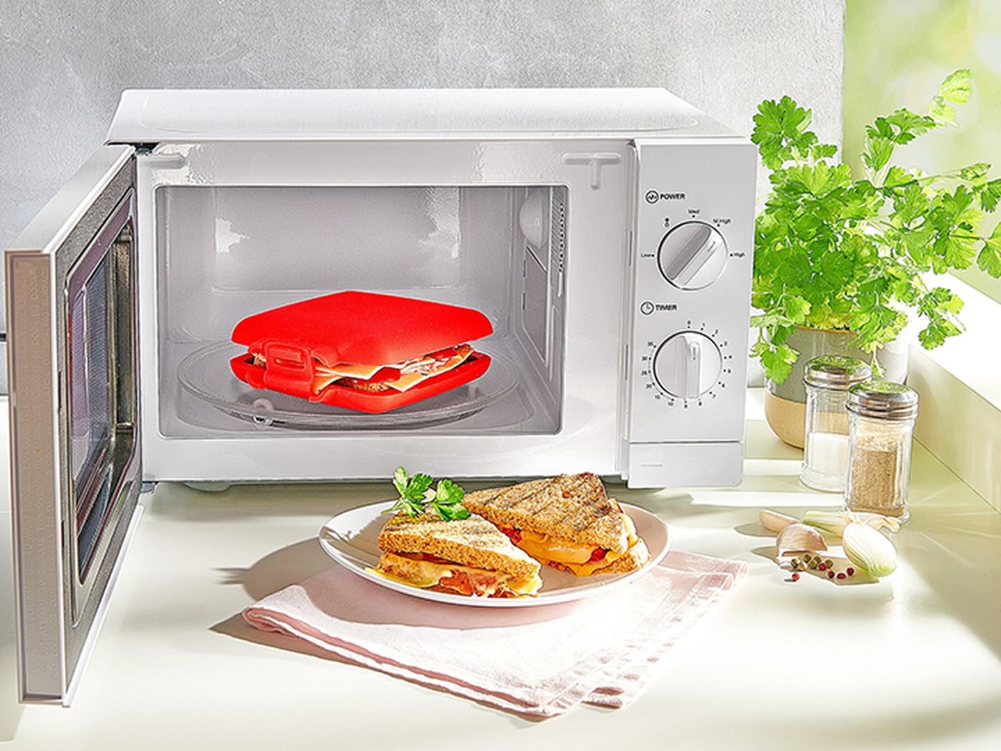 Eine Mikrowelle mit einem roten Sandwichmaker darin, ein Teller mit einem gegrillten Sandwich und Küchenkräuter daneben.