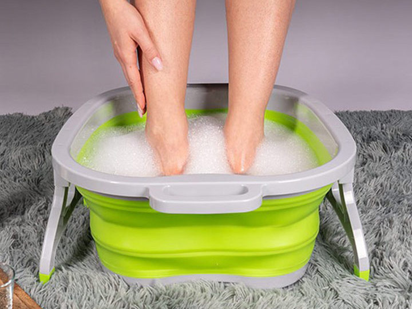 Eine Person weicht ihre Füße in einem grau-grünen, zusammenklappbaren Fußbad ein, das mit Seifenwasser gefüllt ist und auf einem weichen grauen Teppich steht.