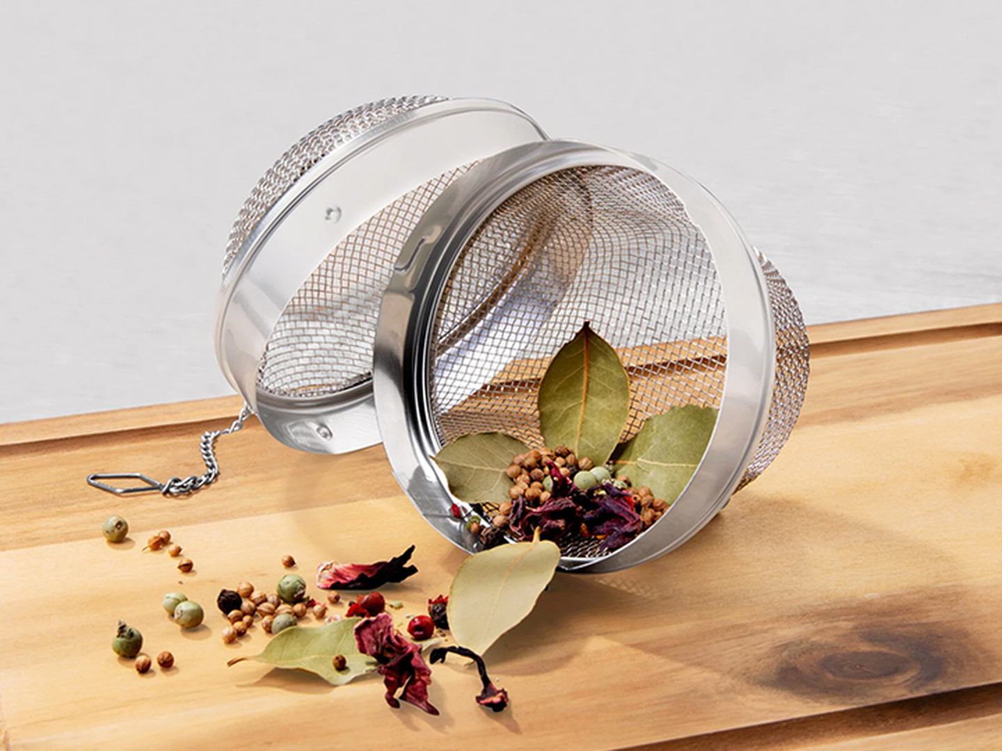 Ein Gewürzsieb aus Metallgeflecht mit Deckel, gefüllt mit getrockneten Kräutern und Gewürzen, liegt geöffnet auf einer Holzoberfläche.