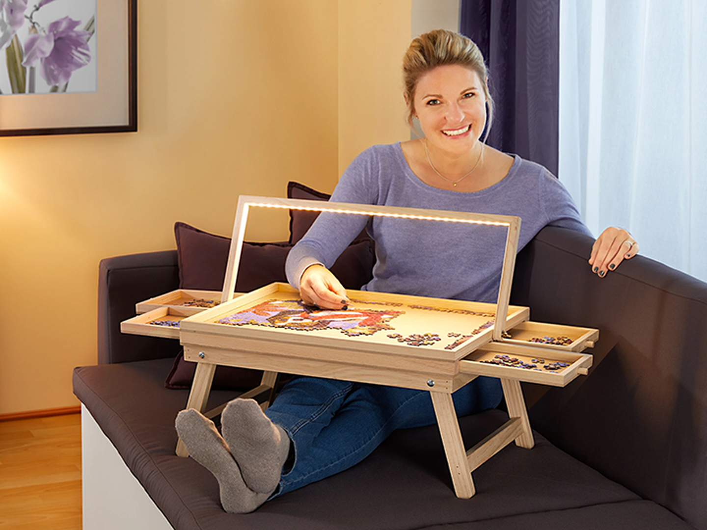 Eine lächelnde Frau setzt bequem auf einem Sofa sitzend ein Puzzle auf einem Puzzletisch mit integrierter Beleuchtung zusammen.