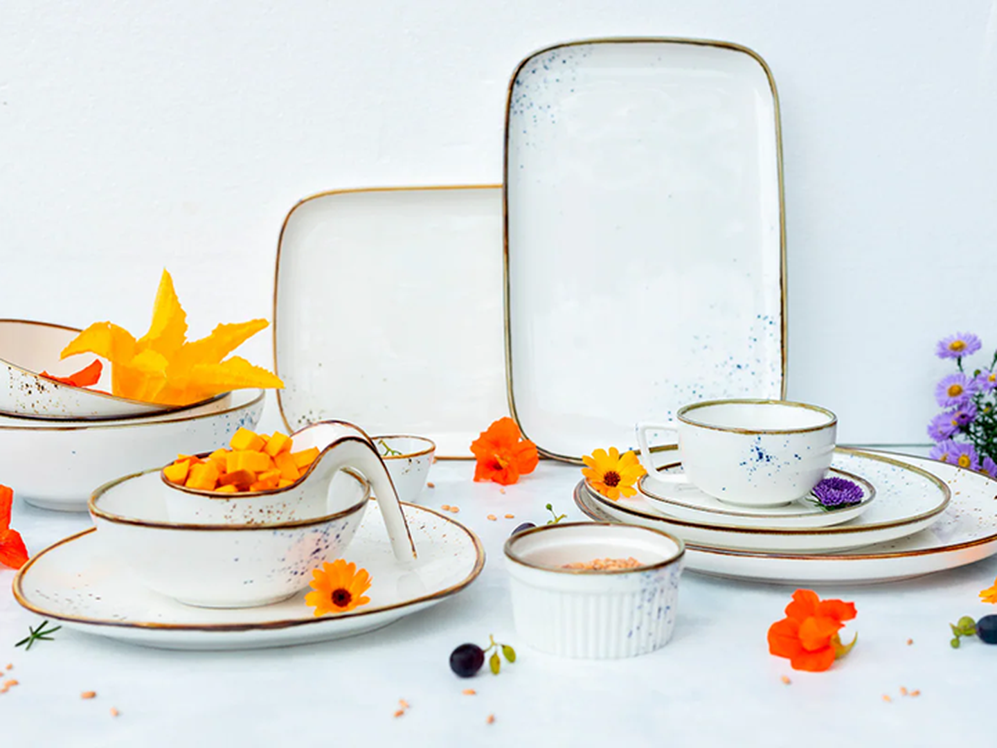 Elegantes weißes Keramik-Essgeschirr-Set mit dezenten Sprenkeln, verziert mit bunten Blumen und einer kleinen Schale mit Speisen, auf hellem Hintergrund.