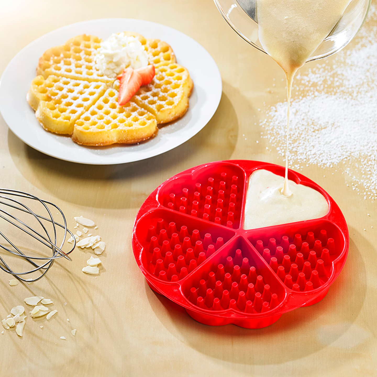 Eine rote Silikon-Waffelform wird mit Teig gefüllt. Ganz in der Nähe liegt eine fertig gebackene, blumenförmige Waffel mit Schlagsahne und Erdbeeren auf einem Teller.
