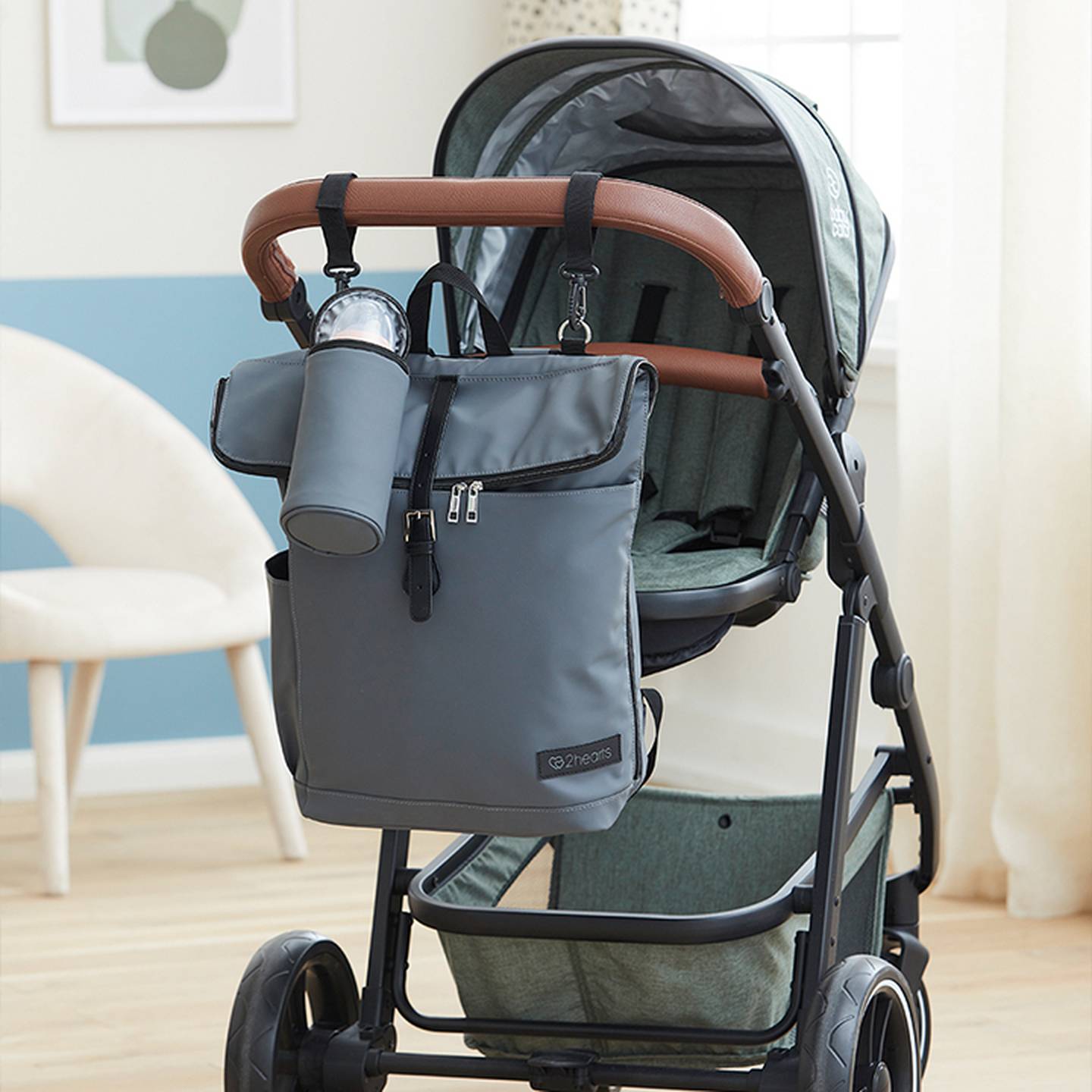 Ein hellgrauer Kinderwagen mit braunen Griffen, an dessen Schiebegriff eine graue Tasche und ein Flaschenhalter befestigt sind. Im Hintergrund sind ein Stuhl und ein Fenster zu sehen.