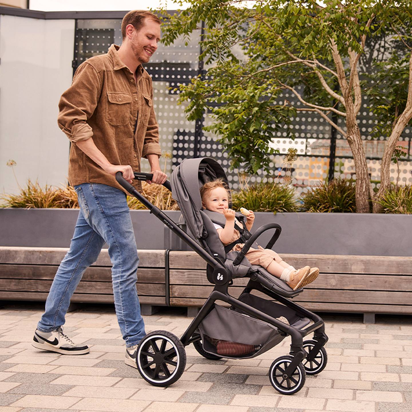 Ein Mann in einer braunen Jacke schiebt ein Kleinkind in einem grauen Kinderwagen einen gepflasterten Weg entlang; im Hintergrund sind Grünanlagen und Bänke zu sehen.