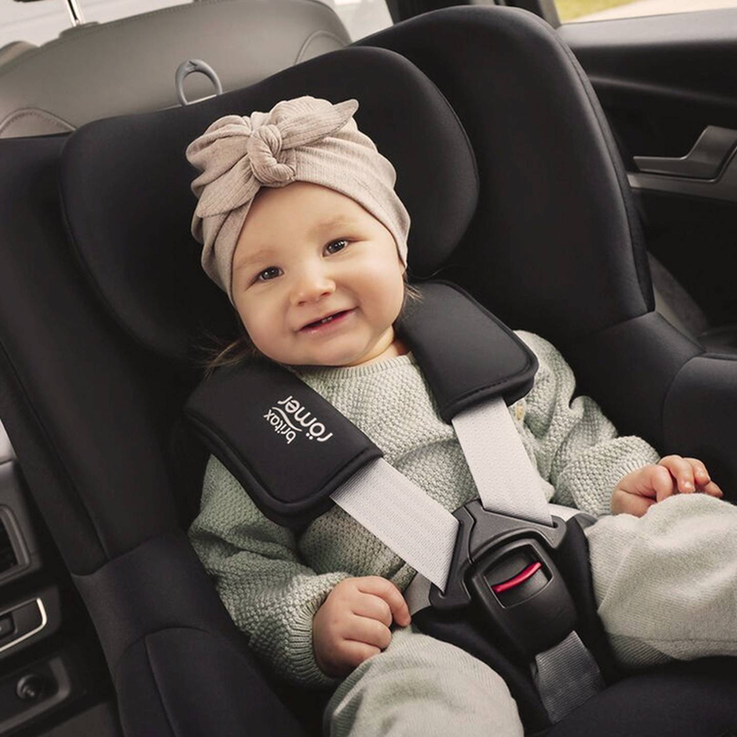 Ein lächelndes Baby mit beigefarbenem Stirnband sitzt in einem Autositz mit schwarzer Polsterung und Sicherheitsgurt.