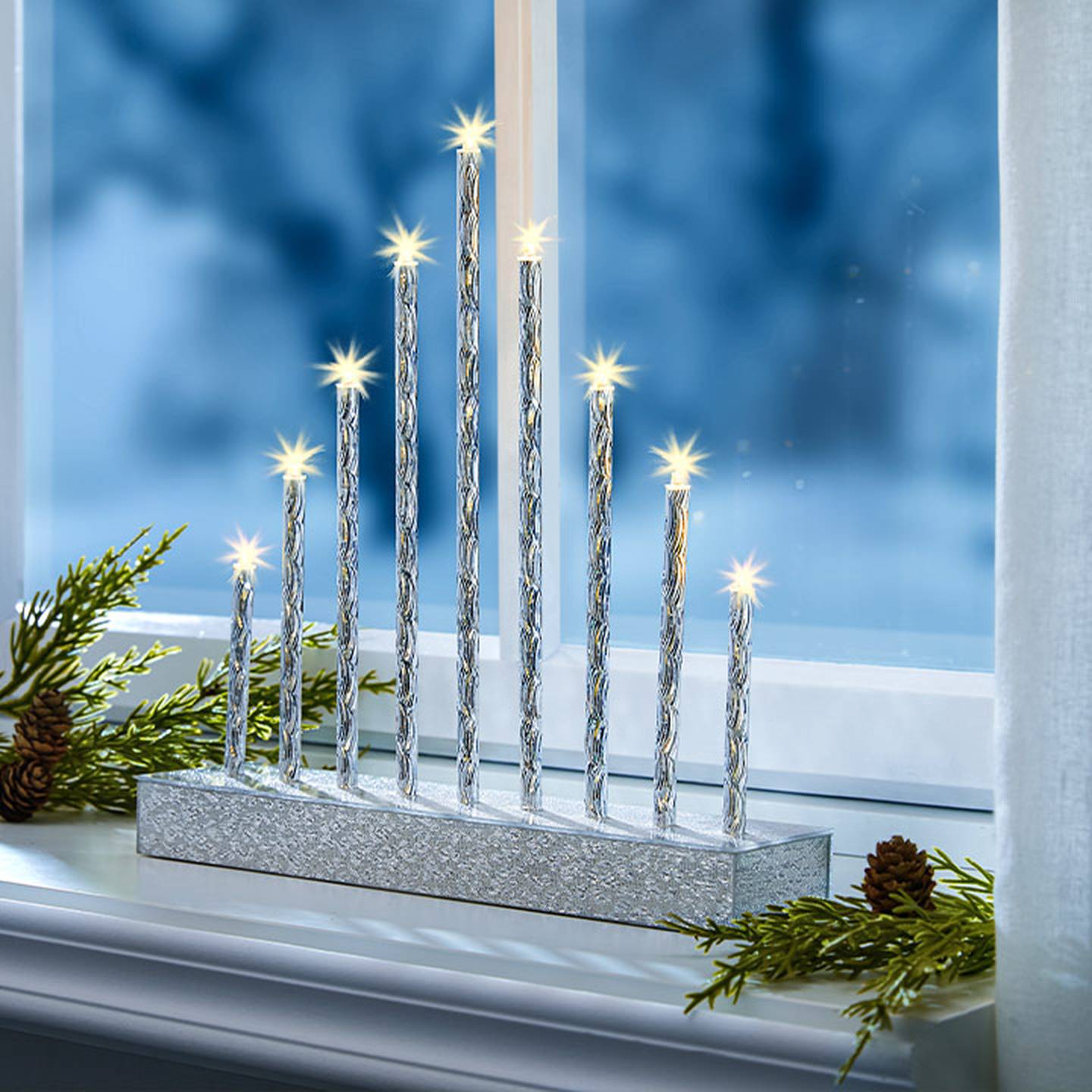 Eine silberne Menora mit brennenden Kerzen auf einem Fensterbrett, verziert mit Kiefernzweigen und Zapfen, vor einem frostig-blauen Fensterhintergrund.