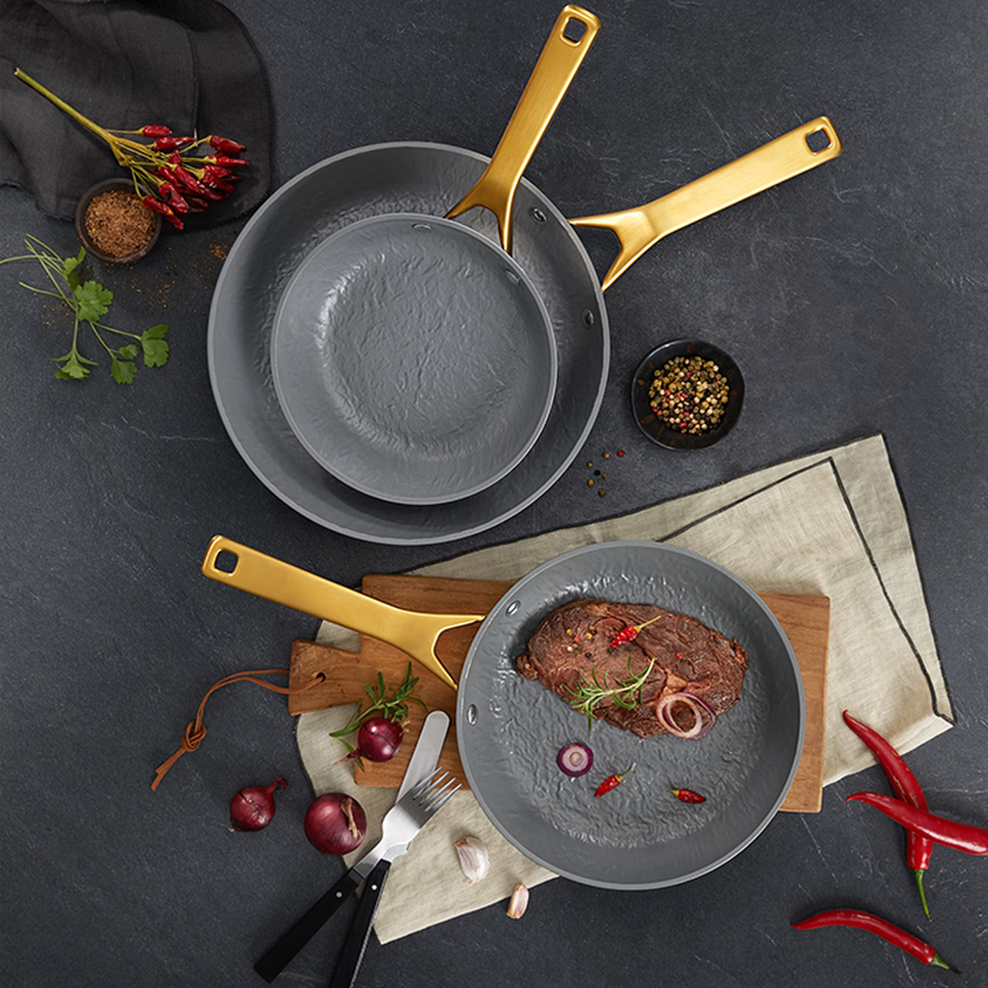 Drei graue Pfannen mit goldenen Griffen auf einer dunklen Oberfläche; eine davon enthält ein gebratenes Steak mit Zwiebeln und Kräutern, umgeben von Paprika und Gewürzen.