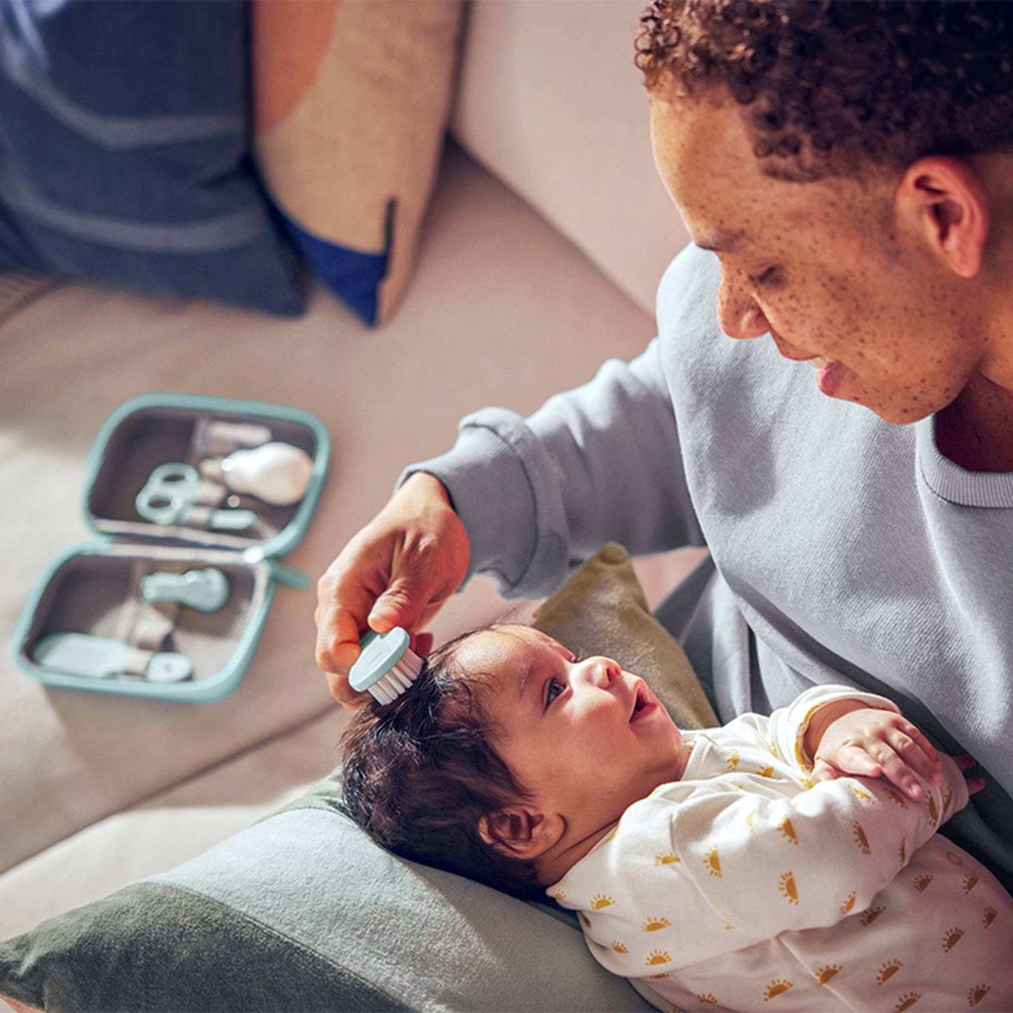 Eine lächelnde Person bürstet sanft die Haare eines Babys mit einer kleinen Bürste. Das Baby liegt behaglich in ihren Armen. Ein Babypflegeset ist in der Nähe zu sehen.