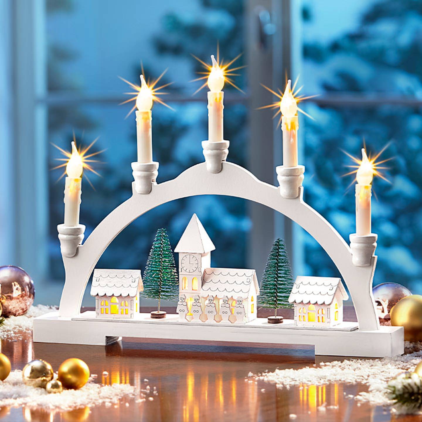 Ein festlicher Kerzenbogen mit brennenden Kerzen, Miniaturhäusern, Bäumen und einem Glockenturm, aufgestellt auf einem Holztisch mit Schnee und Ornamenten.