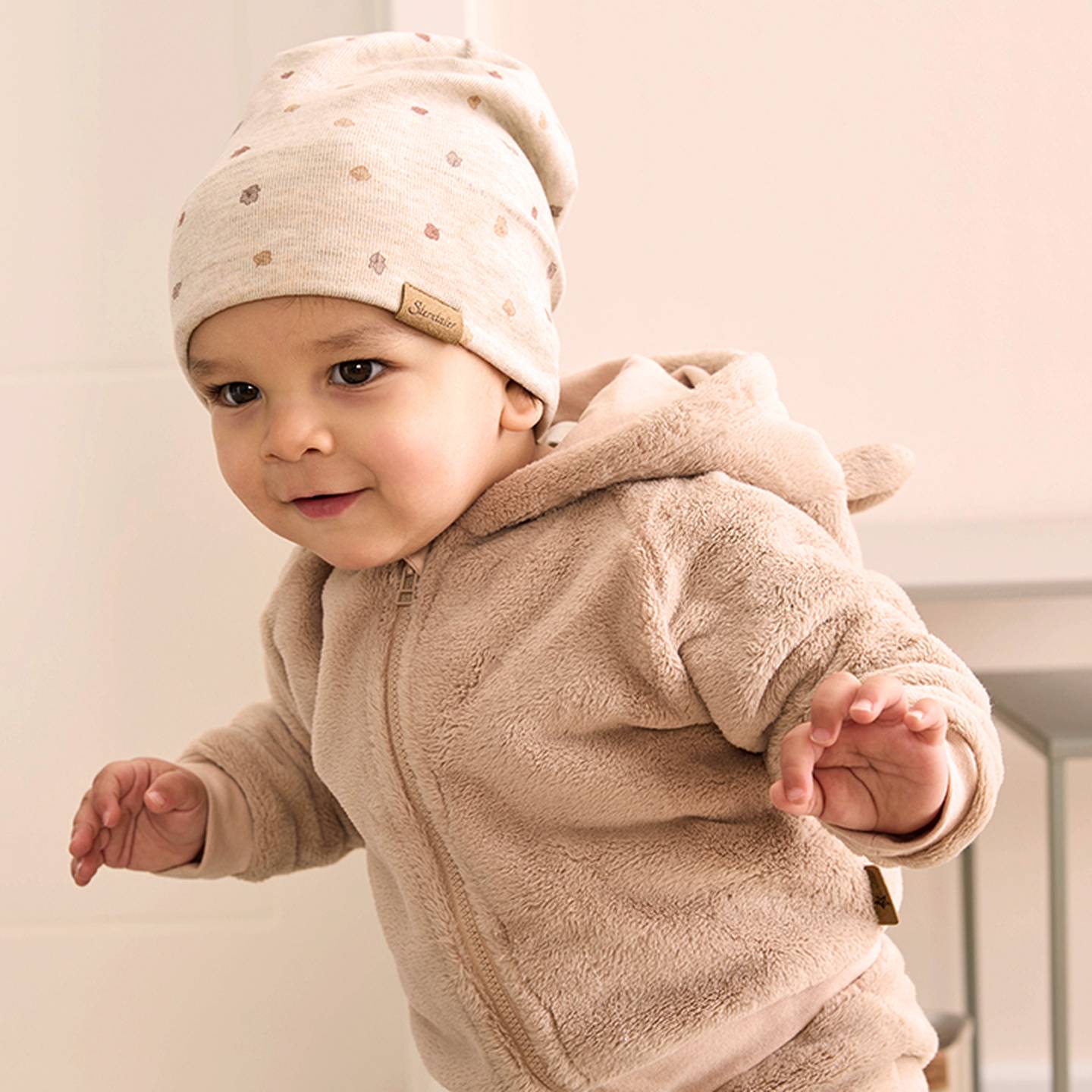 Ein Baby, das einen beigen, flauschigen Kapuzenpullover und eine dazu passende Mütze mit kleinen Mustern trägt, steht mit einem verspielten Gesichtsausdruck drinnen.
