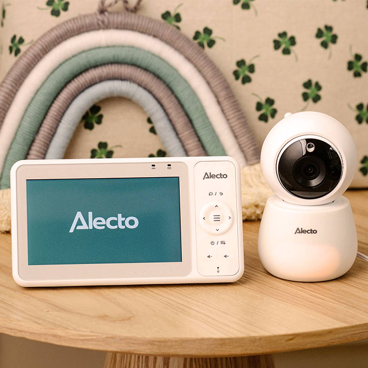 Auf einem Holztisch steht ein weißes Babyphone mit einem Bildschirm, auf dem „Alecto“ steht, und einer Kamera. Im Hintergrund ist ein dekorativer Regenbogen zu sehen.