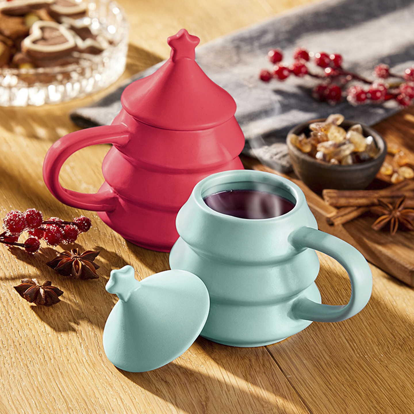 Zwei bunte Tassen mit sternförmigen Deckeln, eine rote und eine blaugrüne, auf einem Holztisch mit Gewürzen, Trockenfrüchten und Keksen im Hintergrund.