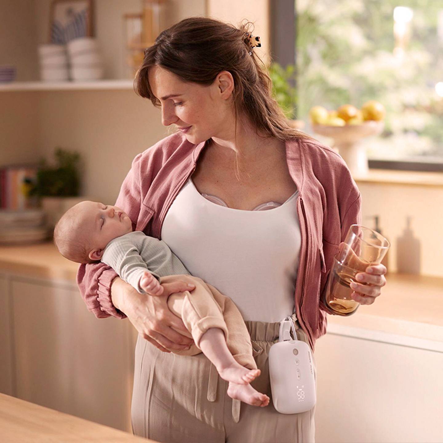 Eine Frau, die ein schlafendes Baby im Arm hält, eine rosa Jacke und eine beige Hose trägt, ein Glas in der Hand hält und eine tragbare Milchpumpe an ihrem Hosenbund befestigt hat.