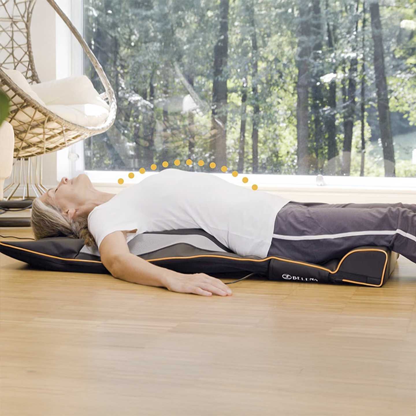 Person, die auf einer Massagematte in einem sonnendurchfluteten Raum mit großen Fenstern liegt, umgeben von Bäumen. Die Matte hat eine gepunktete Linie, die die Ausrichtung der Körperhaltung anzeigt.