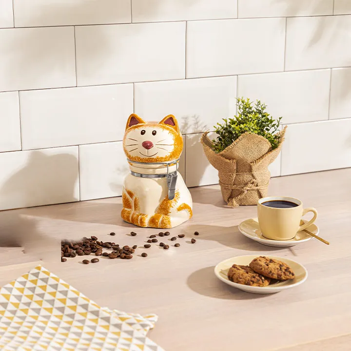 Eine katzenförmige Keksdose auf einer Küchentheke mit Kaffee, Keksen, Kaffeebohnen, einer Topfpflanze und einem gelb gemusterten Tuch in der Nähe.