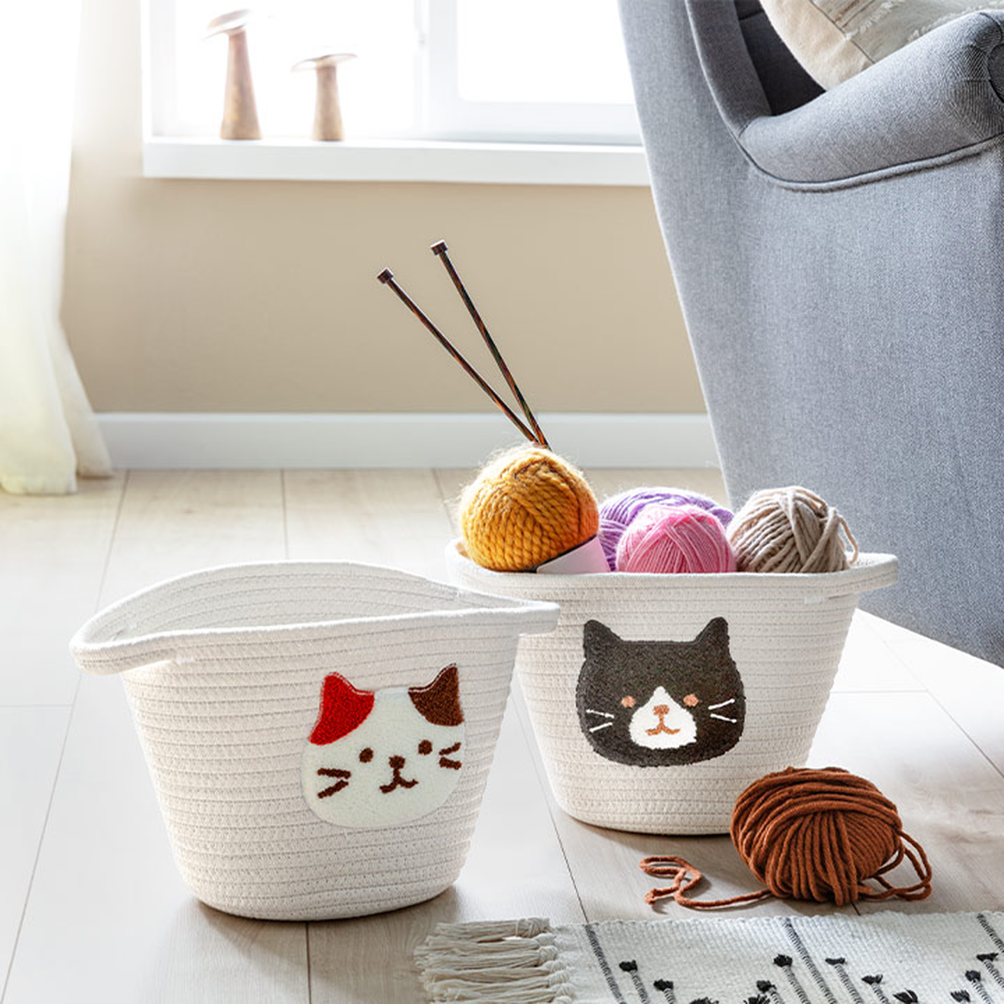 Zwei weiße geflochtene Körbe mit Katzengesichtern, gefüllt mit bunten Wollknäueln und Stricknadeln, stehen auf einem hellen Holzboden neben einem Sofa.