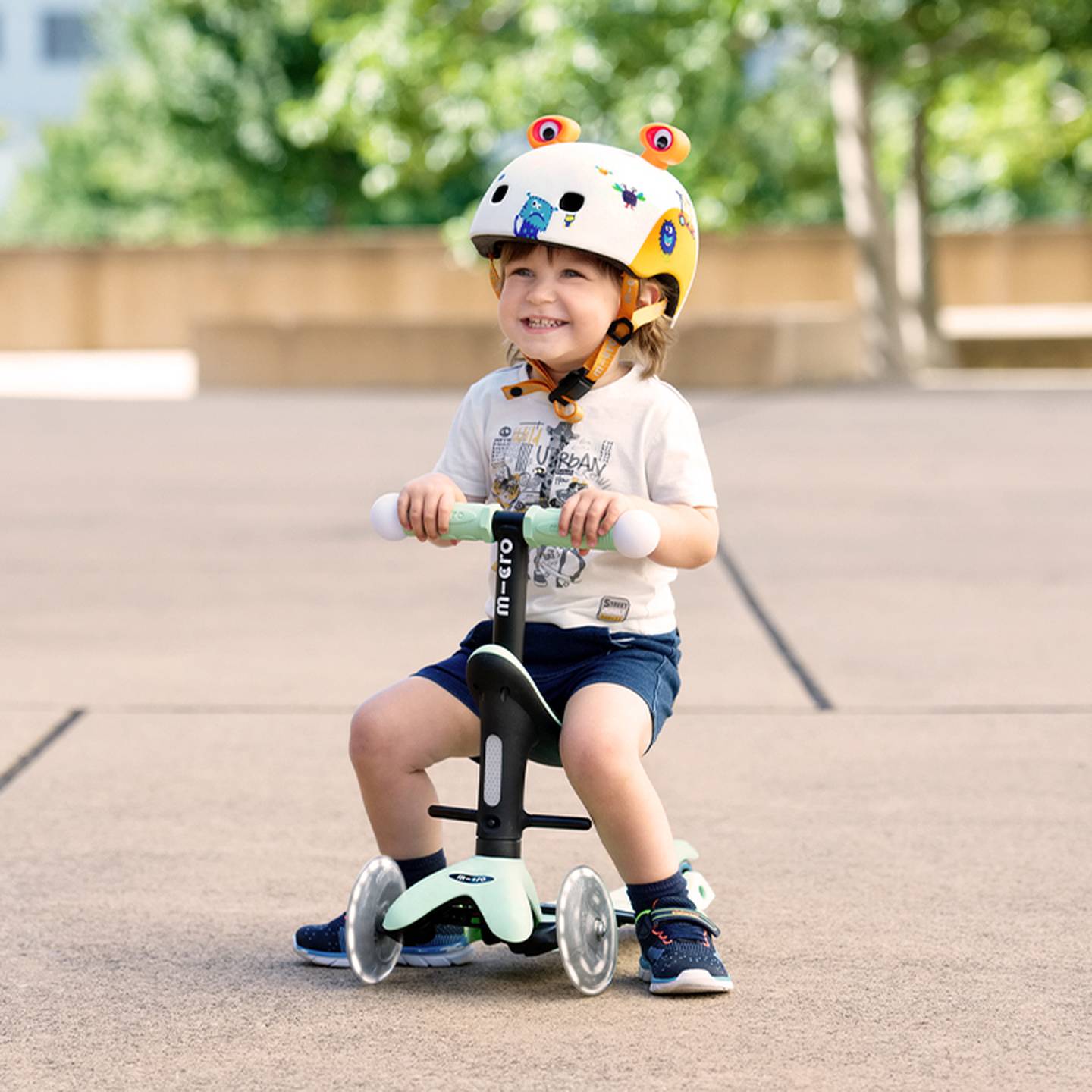 Ein Kind mit einem Helm mit Froschmotiv lächelt, während es auf einem kleinen Roller mit Stützrädern auf einer gepflasterten Fläche fährt, im Hintergrund sind Bäume zu sehen.