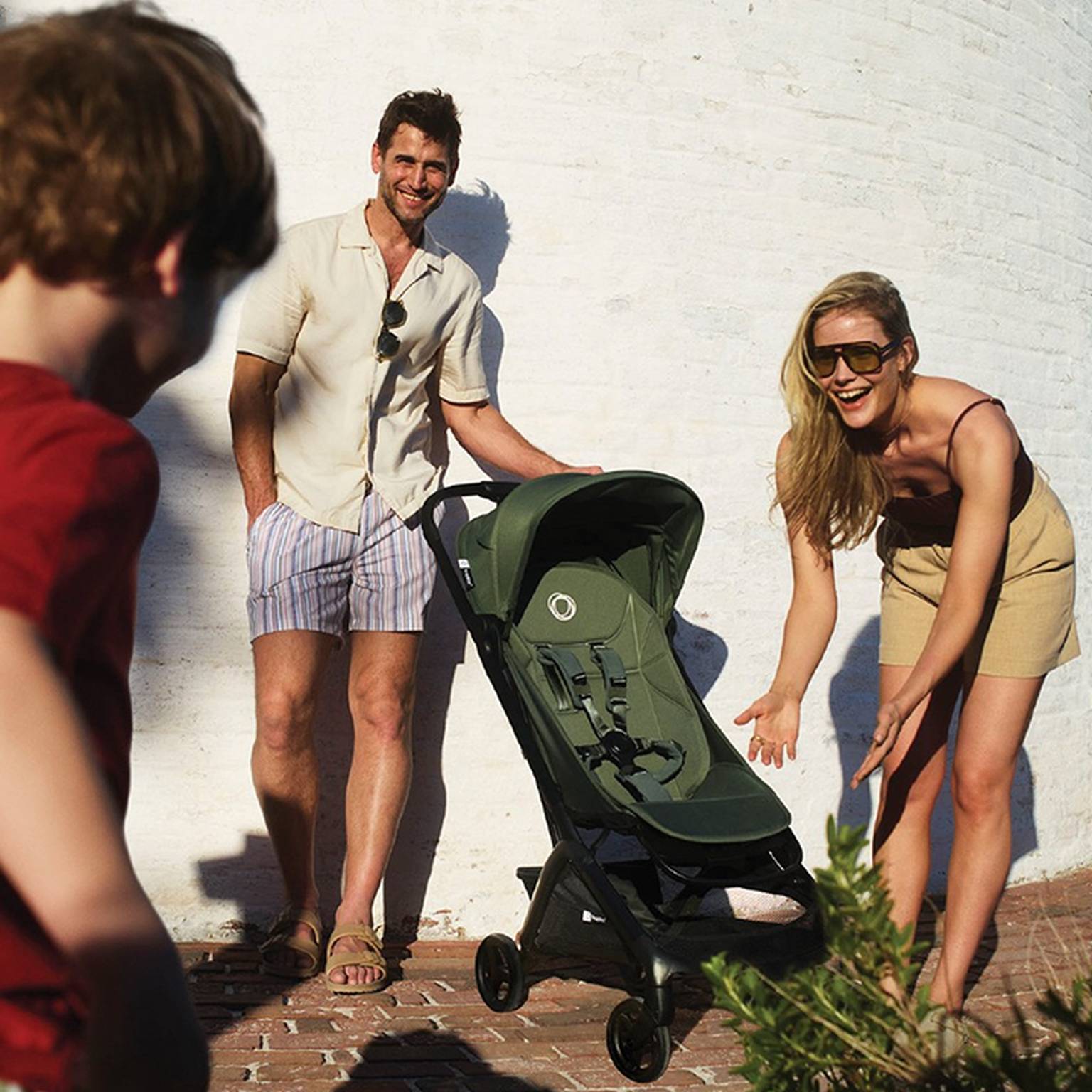 Ein Paar lächelt einem Kind zu und zeigt einen grünen Kinderwagen vor einer weißen Backsteinmauer. Die Frau gestikuliert in Richtung des Kinderwagens.