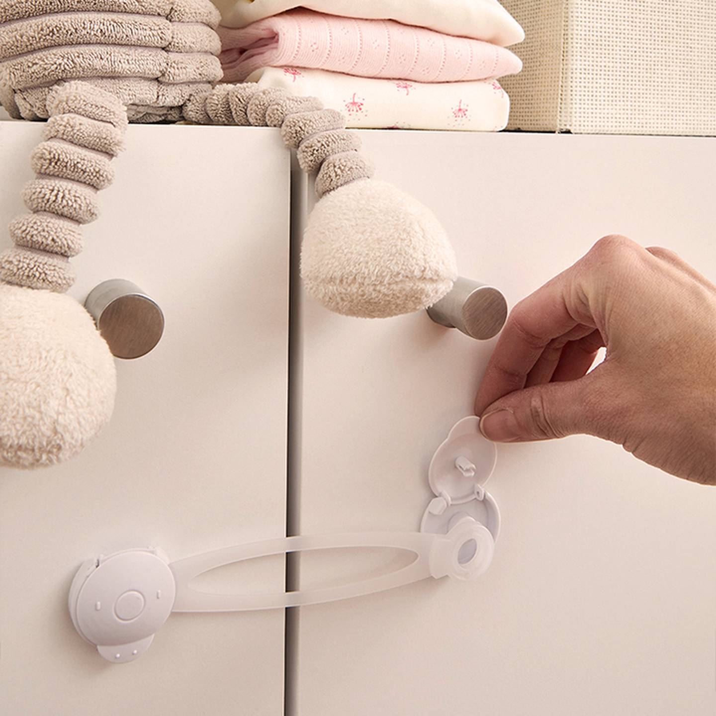 Eine erwachsene Hand öffnet den weißen Kindersicherungsriegel aus Kunststoff an den Schranktüren; darauf liegen ein Stofftier und gefaltete Babykleidung.