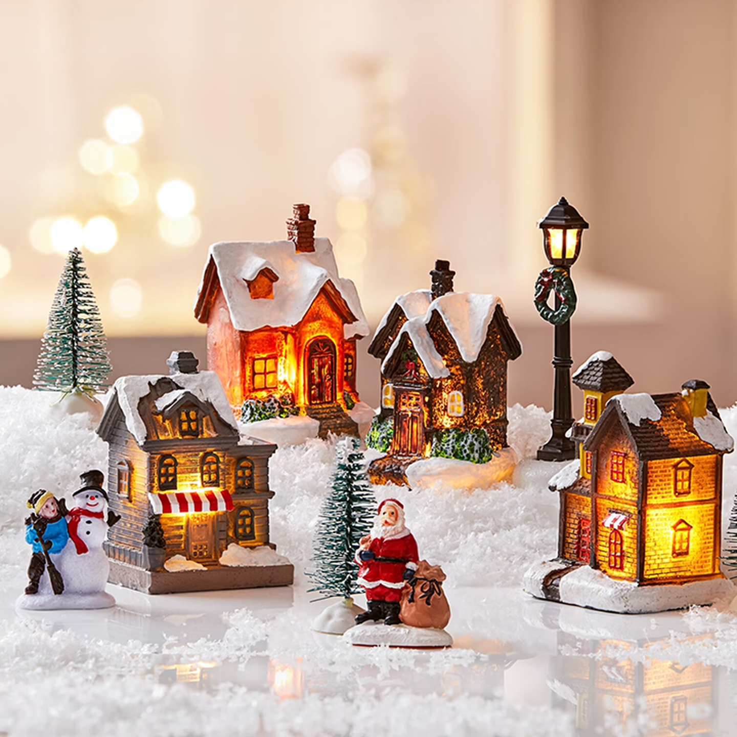 Ein kleines Winterdorf mit 4 Häusern, 2 Menschen und Bäumen mit LEDs umgeben von Kunstschnee