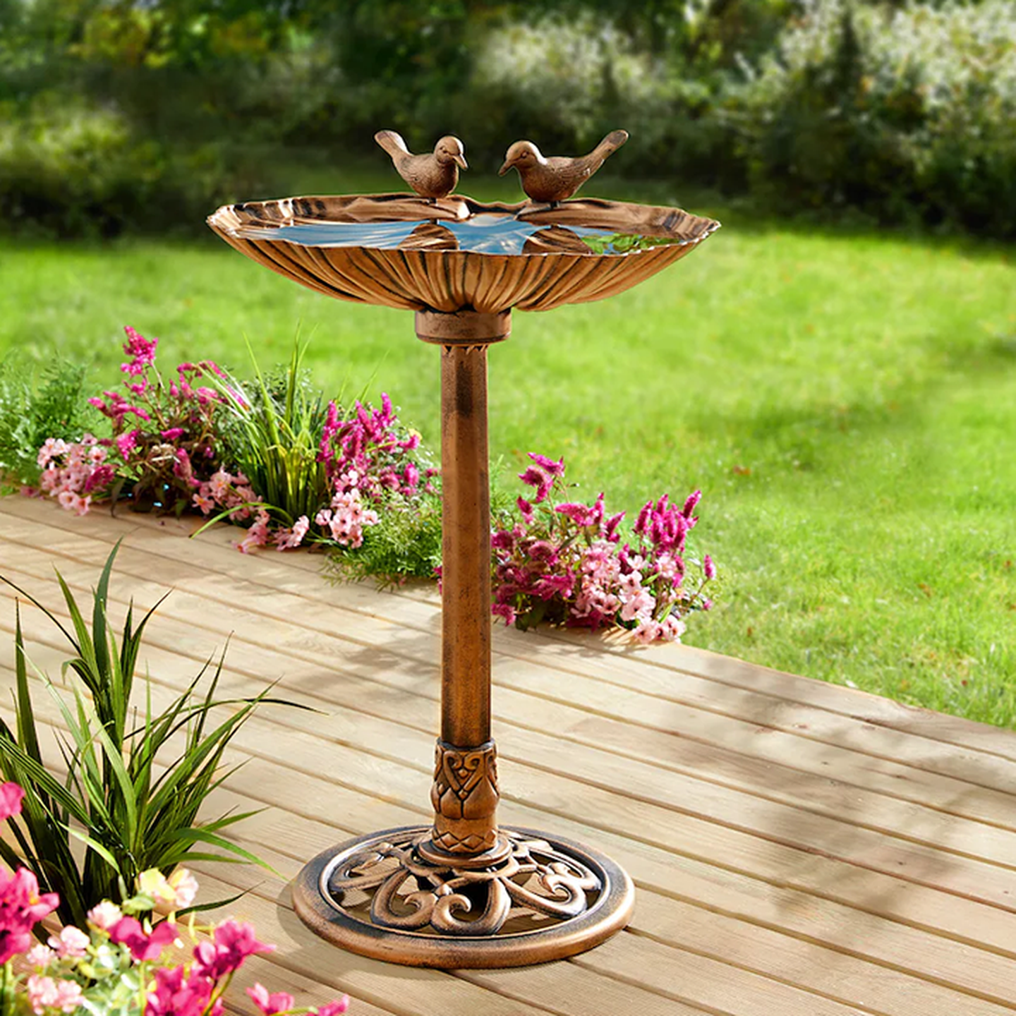 Vogeltränke aus Bronze mit zwei Vogelskulpturen auf einer Holzterrasse, umgeben von rosa Blumen und sattgrünem Gras.