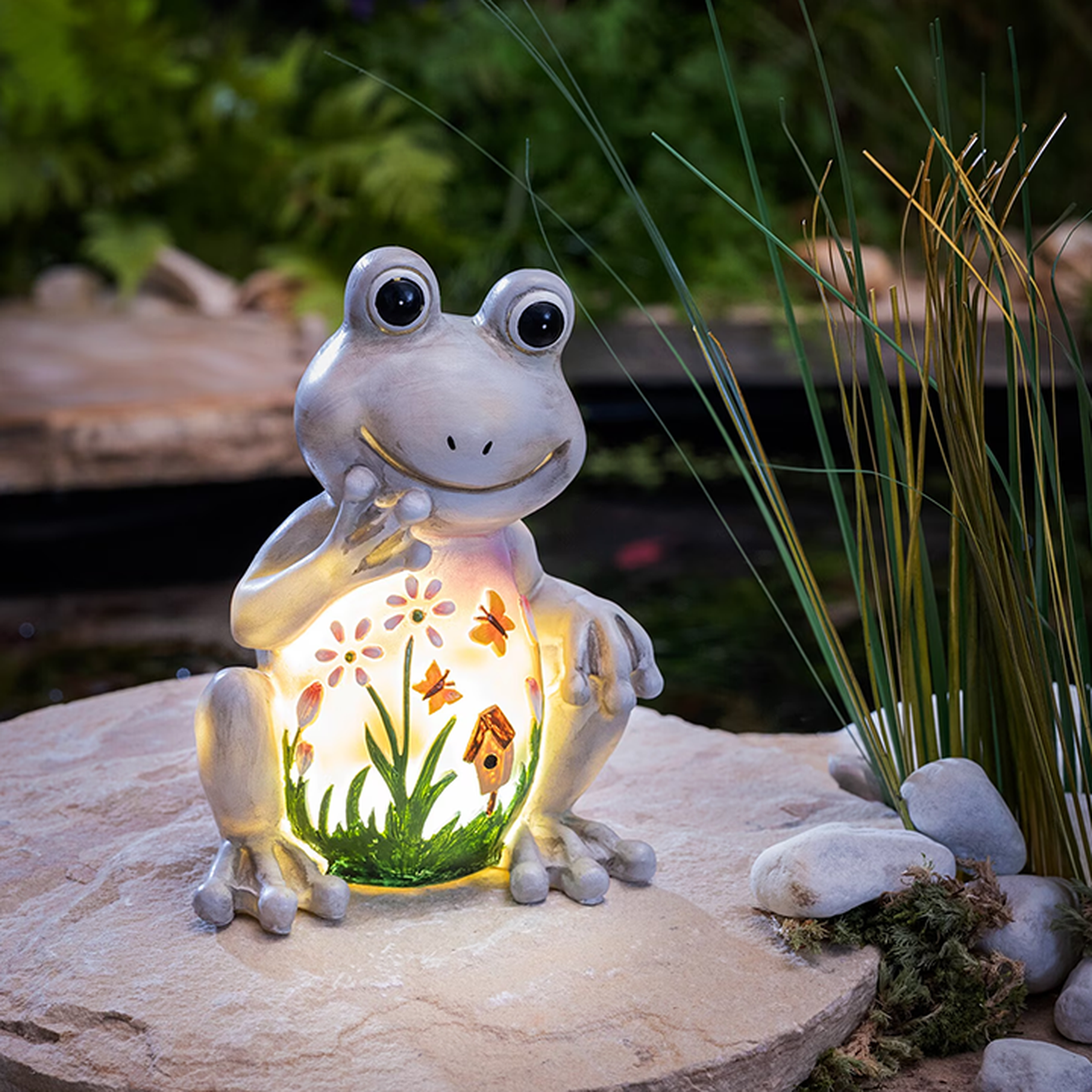 Eine verspielte Froschfigur mit leuchtendem Bauch, verziert mit Blumen, Schmetterlingen und einem Vogelhäuschen, sitzt auf einem Stein im Garten.