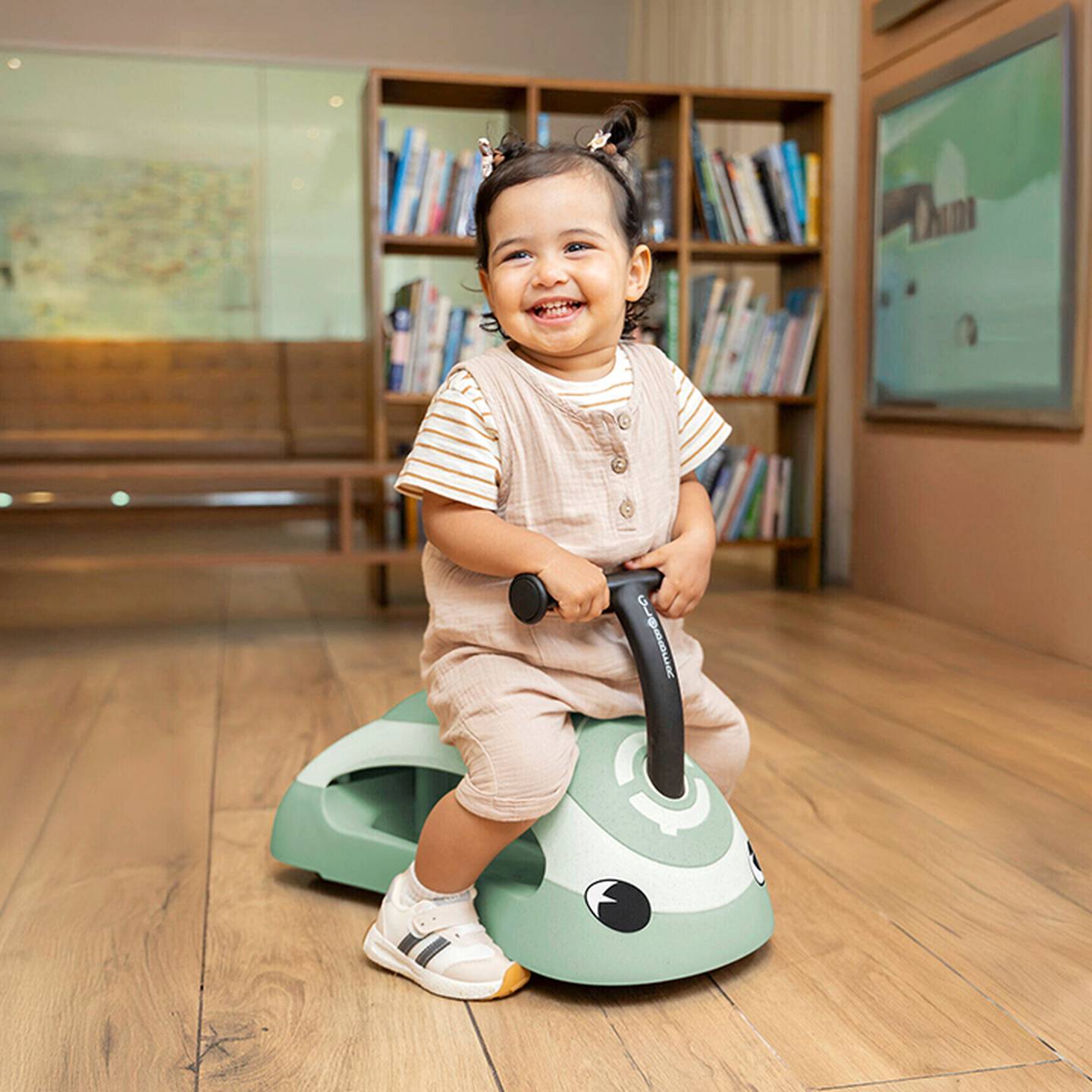 Kleinkind lächelt und sitzt drinnen auf einem grünen Spielzeugroller, im Hintergrund sind Bücherregale und ein Holzboden zu sehen.