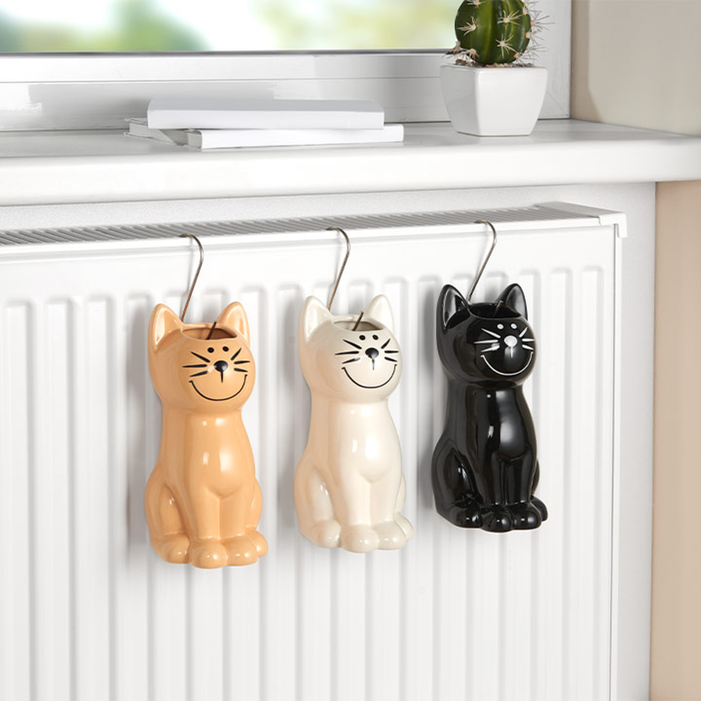 An einem Heizkörper hängen drei Luftbefeuchter aus Keramik in Katzenform in Beige, Weiß und Schwarz. Auf der Fensterbank darüber stehen ein kleiner Kaktus und ein Buch.