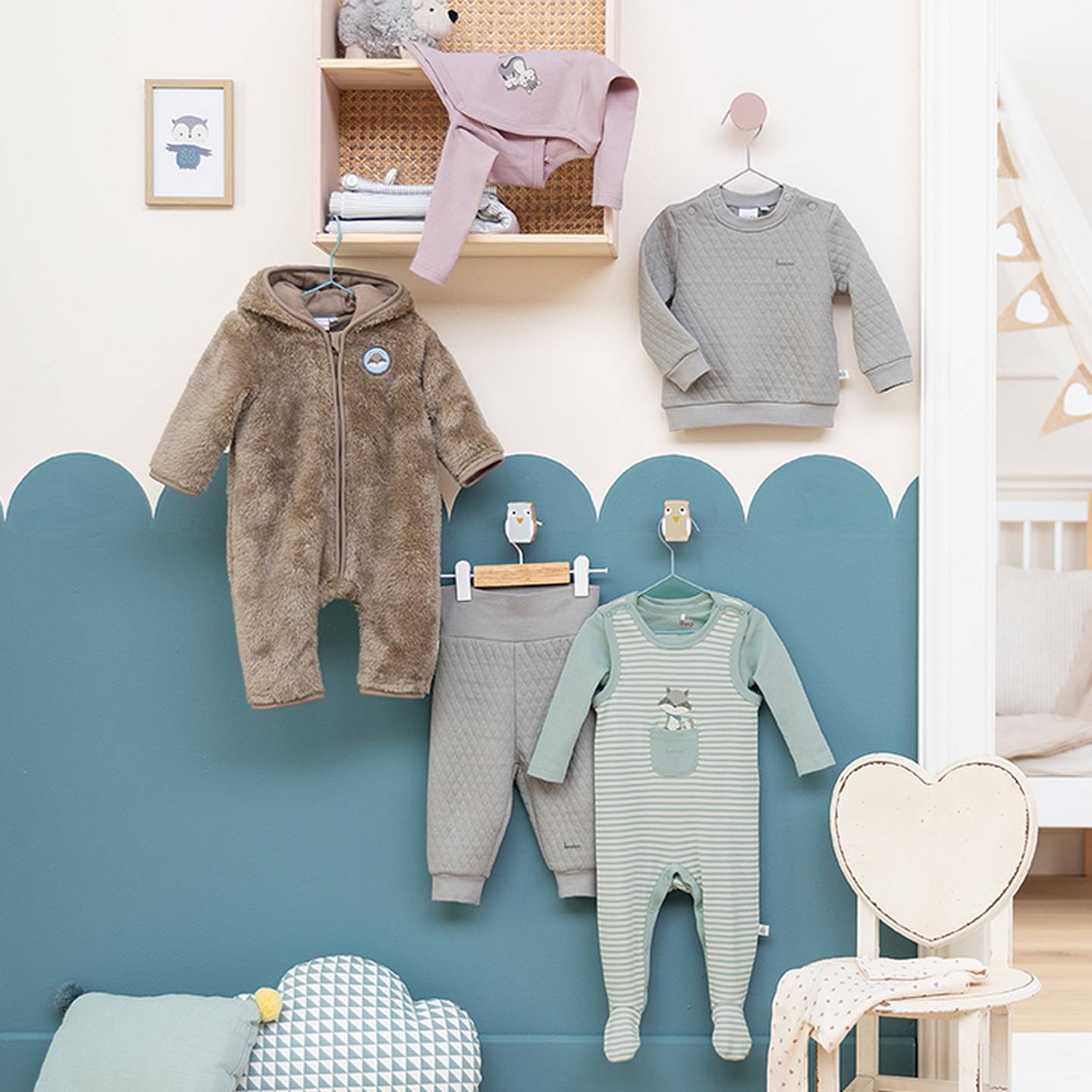 An einer Wand sind Kinderkleidungsstücke ausgestellt, darunter ein brauner Flauschanzug sowie graue und grüne Outfits, darunter ein herzförmiger Stuhl.