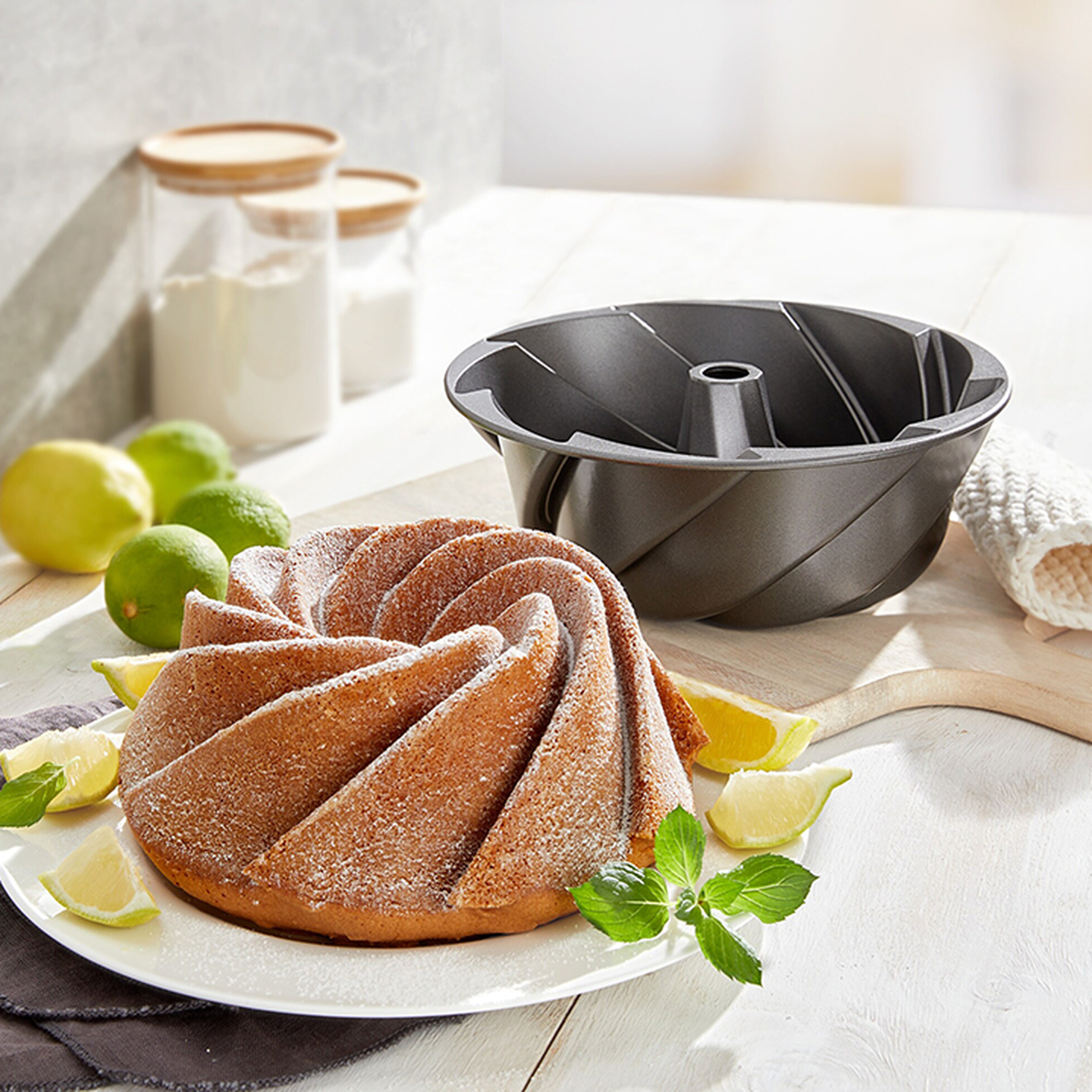 Marmorierter Gugelhupf, mit Puderzucker bestäubt, auf einem Teller; daneben liegen Limetten, ein Minzzweig und die passende Gugelhupfform auf einem Holztisch.