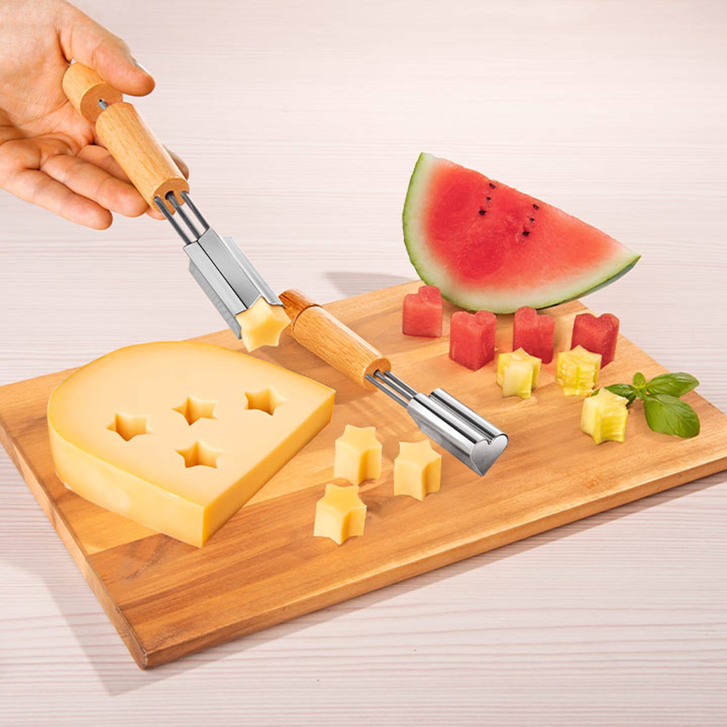 Ein Holzbrett mit sternförmigen Käse- und Wassermelonenstücken, eine Hand, die einen sternförmigen Ausstecher hält, und Basilikum als Garnitur.