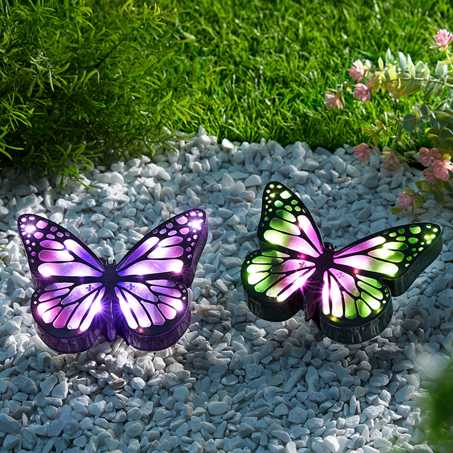 Zwei leuchtende Schmetterlings-Gartenleuchten auf weißen Kieselsteinen, umgeben von grünem Gras und rosa Blumen.