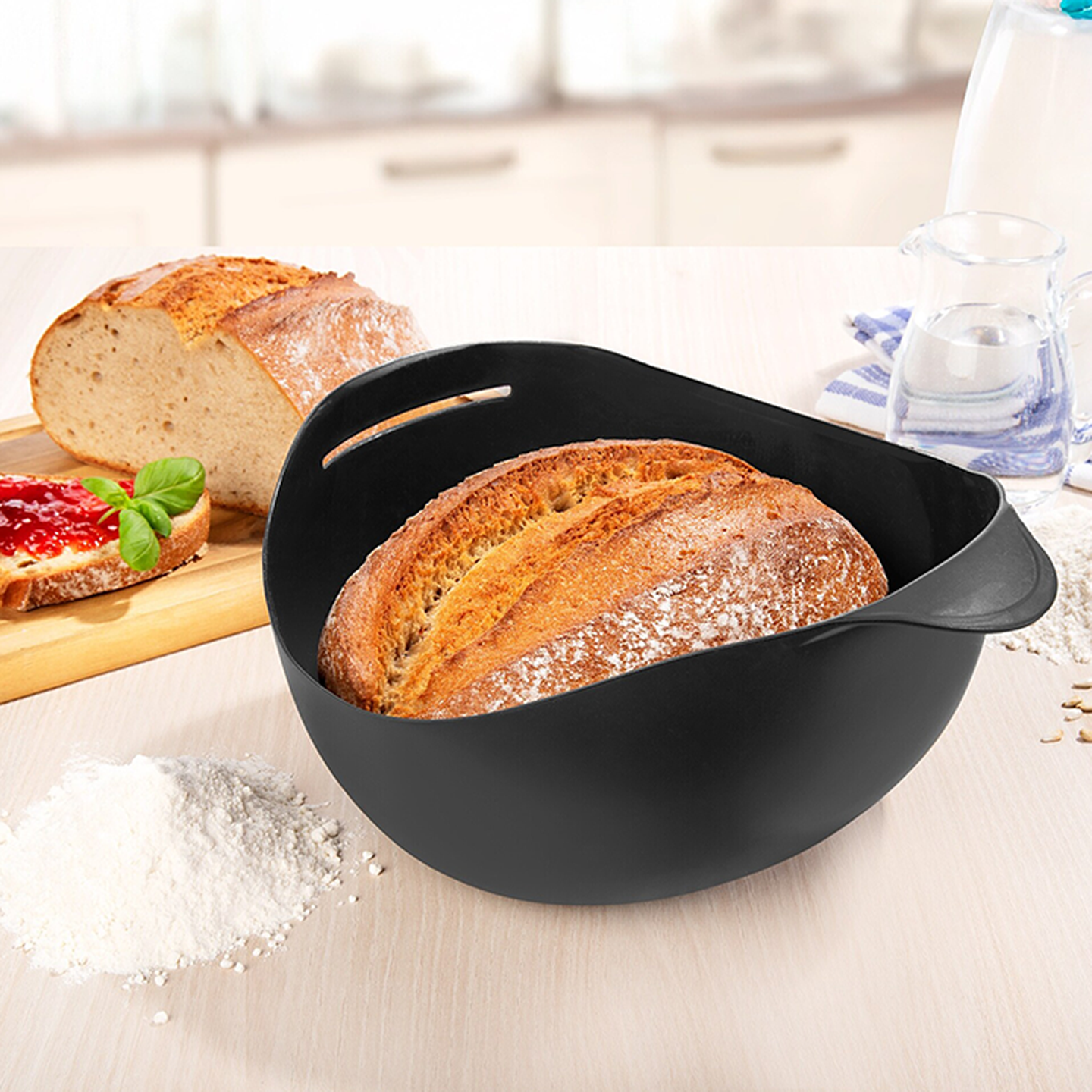 Frisch gebackenes, knuspriges Brot in einer schwarzen Backschale auf einer Küchenarbeitsplatte; daneben liegen Brotscheiben, Mehl und ein Glas Wasser.