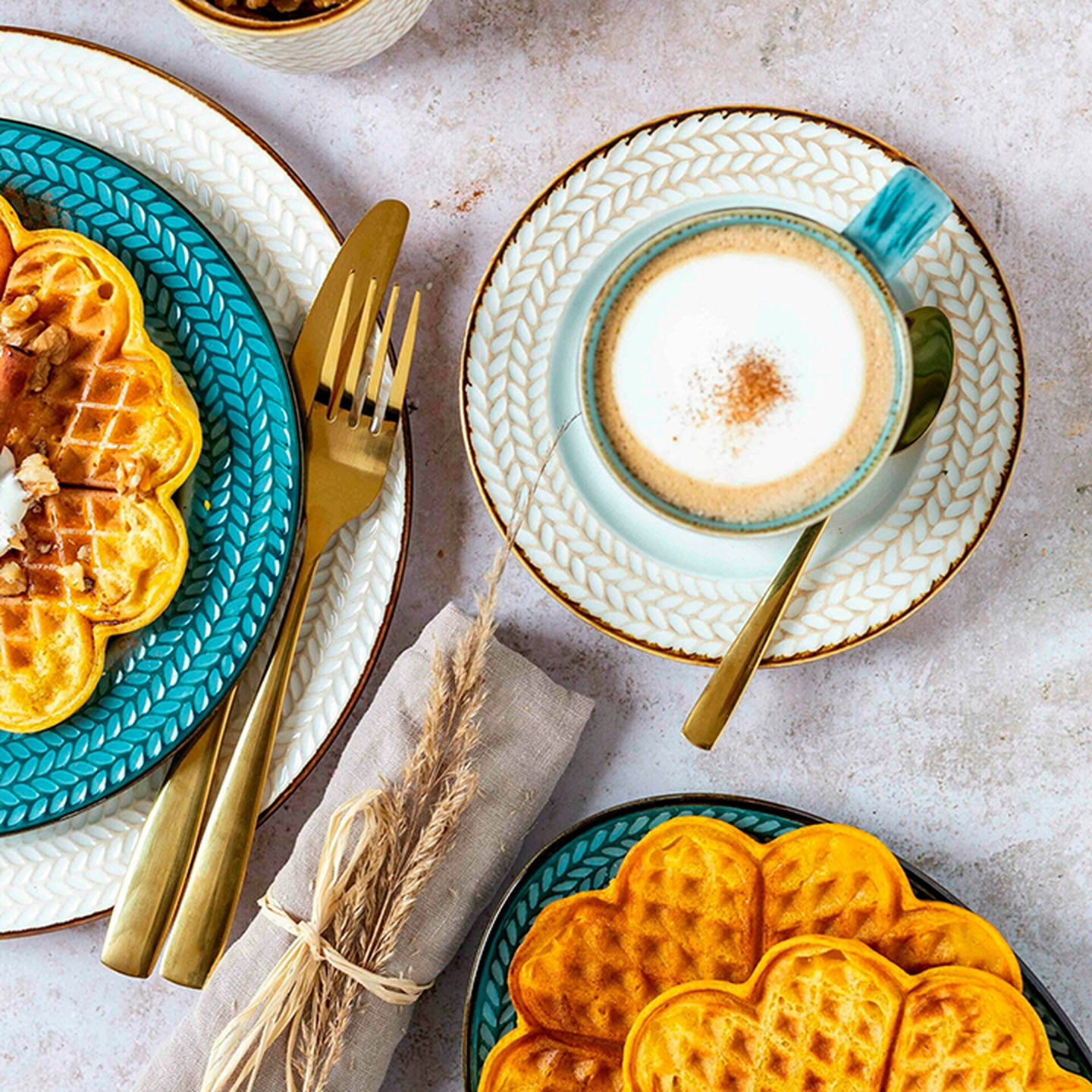 Herzförmige Waffeln auf einem türkisfarbenen Teller mit goldenem Besteck, daneben eine Tasse Cappuccino auf einer weißen Untertasse.