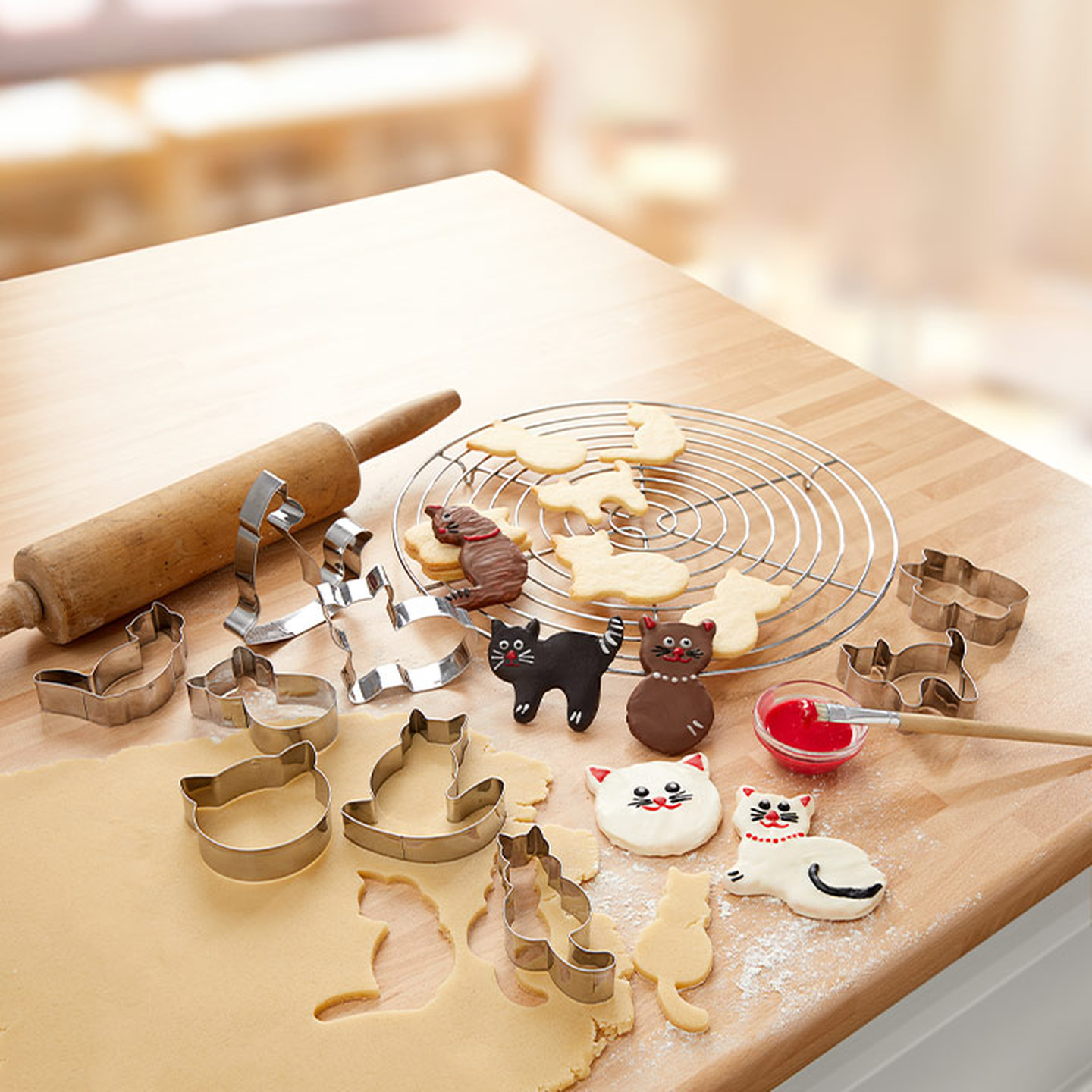 Katzenförmige Ausstechformen, Teig, verzierte Katzenkekse und ein Nudelholz auf einem Holztisch. Kühlgitter mit Keksen im Hintergrund.