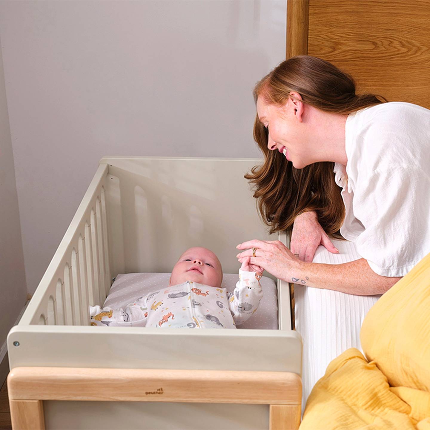 Eine Frau lächelt, während sie sanft die Hand eines Babys hält, das in einem Kinderbett liegt – ein warmherziger und zärtlicher Moment.