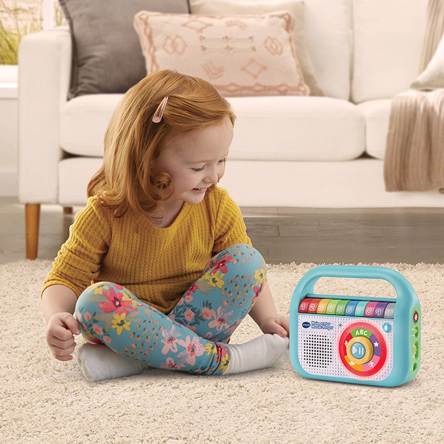 Ein Kind mit roten Haaren, das ein gelbes Oberteil und Leggings mit Blumenmuster trägt, sitzt auf einem Teppich und spielt mit einem bunten Lernspielzeugradio.