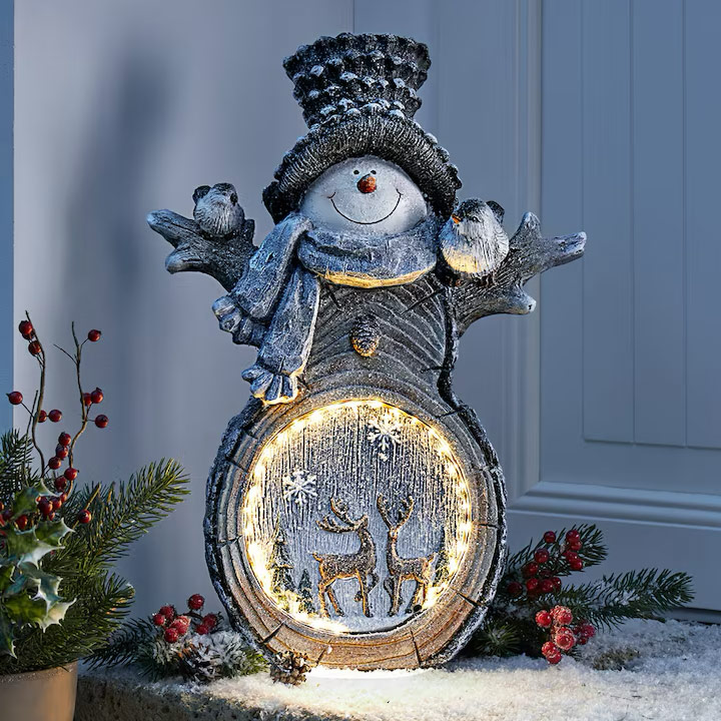 Festliche Schneemannstatue mit Hut und Schal, Vögeln auf den Armen, leuchtendes kreisförmiges Paneel mit zwei Rentieren, umgeben von Stechpalmenzweigen und Schnee.