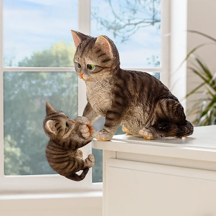 Zwei realistische Katzenskulpturen: eine erwachsene Katze und ein Kätzchen. Die erwachsene Katze scheint dem Kätzchen auf eine weiße Fläche zu helfen. Im Hintergrund ist ein Fenster zu sehen.