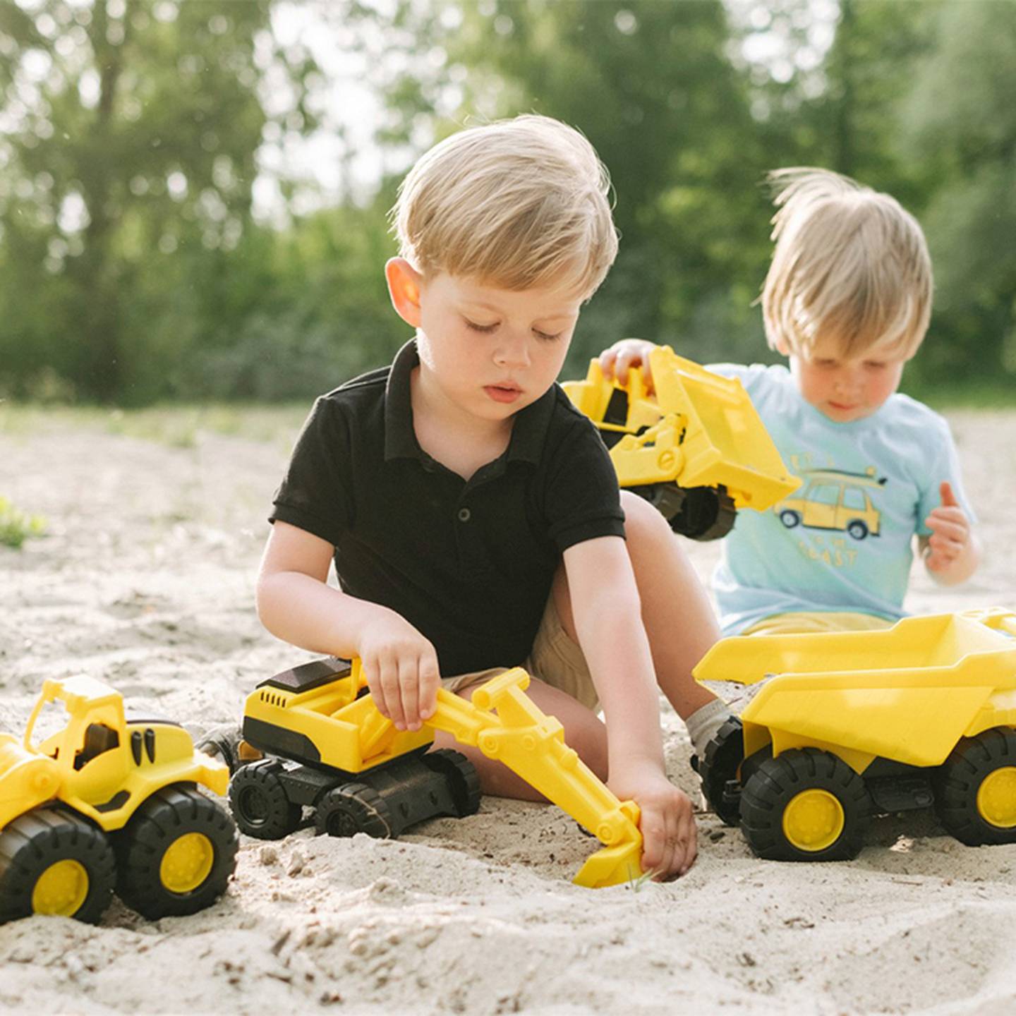 Zwei kleine Kinder spielen im Sand, umgeben von Grün, mit gelben Spielzeug-Baufahrzeugen.