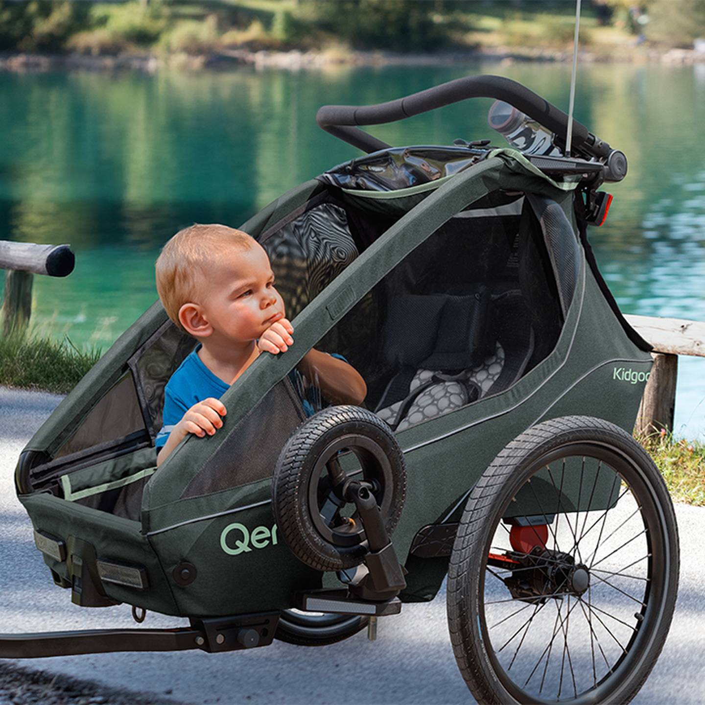 Ein Kleinkind sitzt nachdenklich in einem grünen Fahrradanhänger. Im Hintergrund sieht man einen ruhigen See und viel Grün.