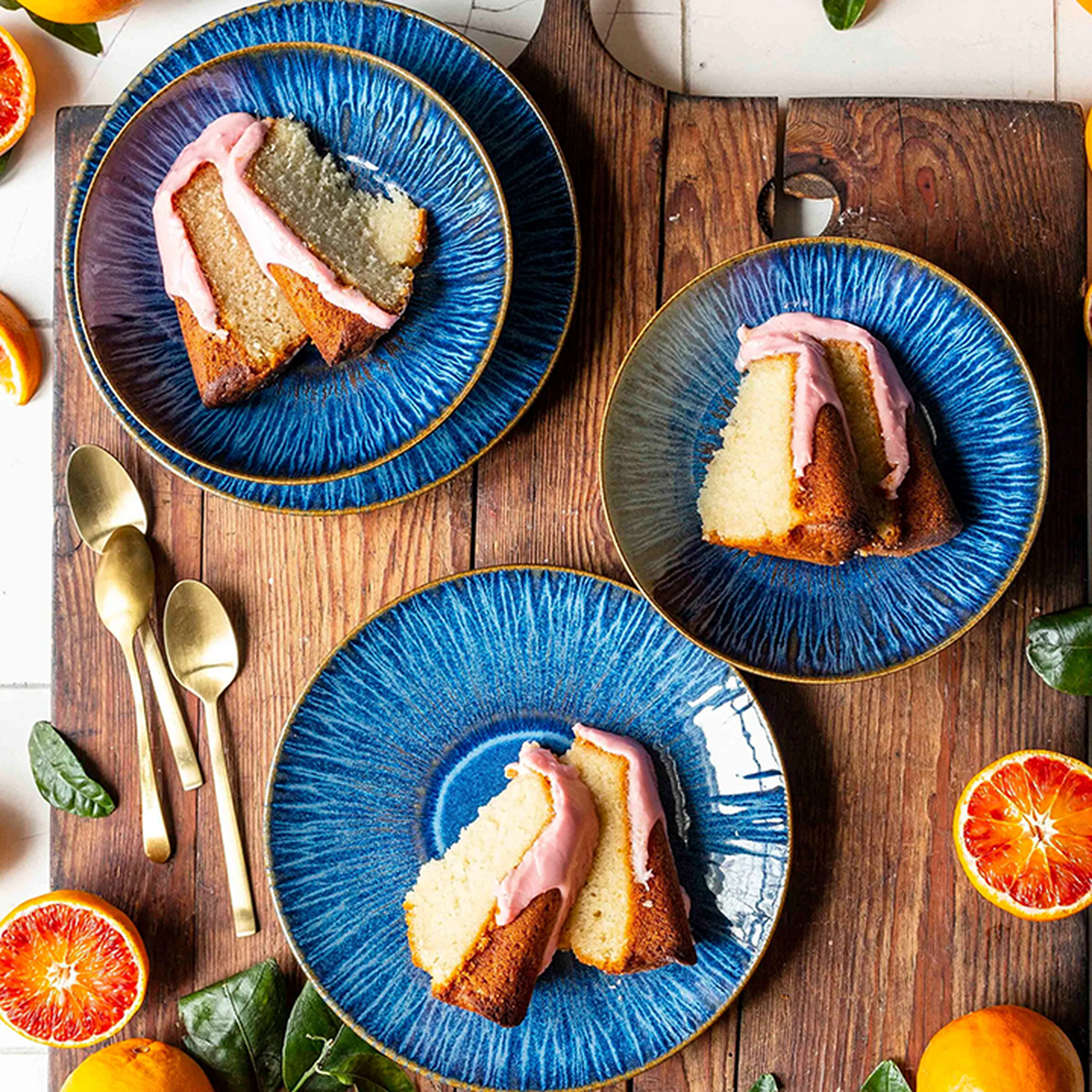 Drei Kuchenstücke mit rosa Glasur auf blauen Tellern, angerichtet auf einem Holzbrett, umgeben von goldenen Löffeln und Orangenscheiben.