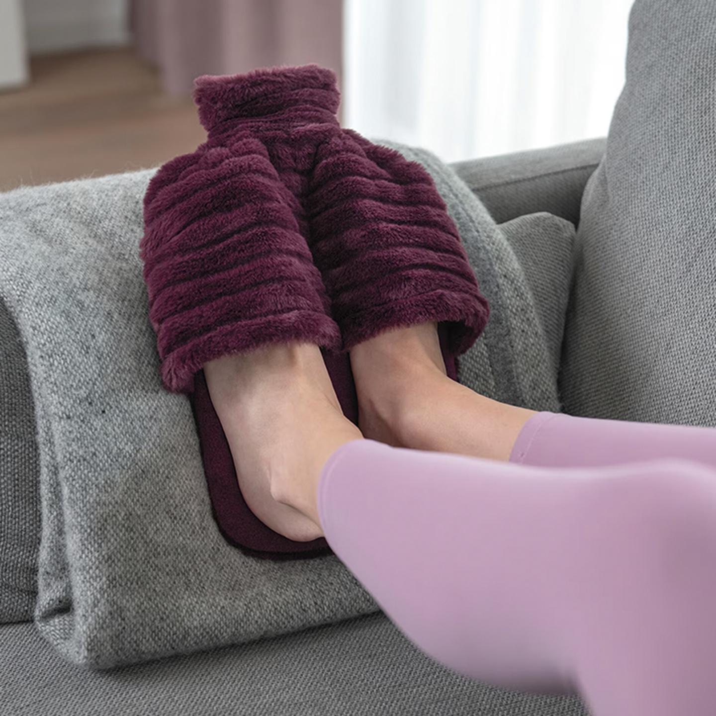 Füße in rosa Leggings ruhen in weichen, burgunderroten Hausschuhen auf einer grauen Decke, die über einer Couch liegt.