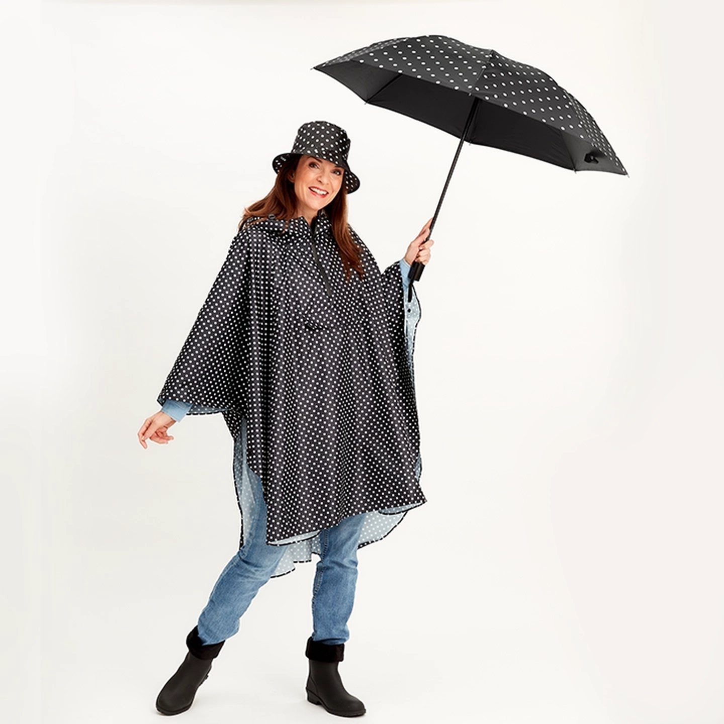 Eine lächelnde Frau in einem gepunkteten Regenmantel und Hut, die einen passenden Regenschirm hält, steht auf einem weißen Hintergrund.