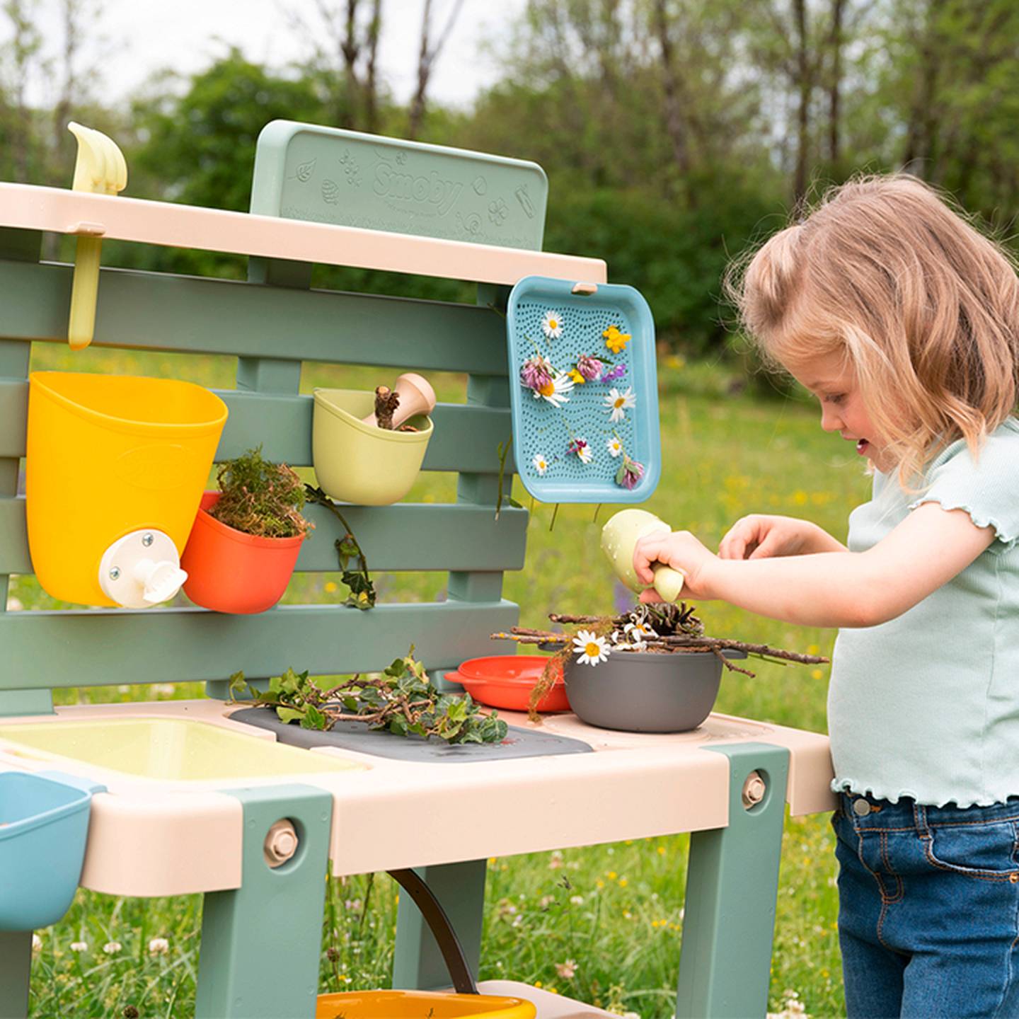 Ein Kind spielt im Freien mit einer Spielküche, arrangiert Blumen und Blätter in Behältern, umgeben von bunten Plastikbehältern und -utensilien.