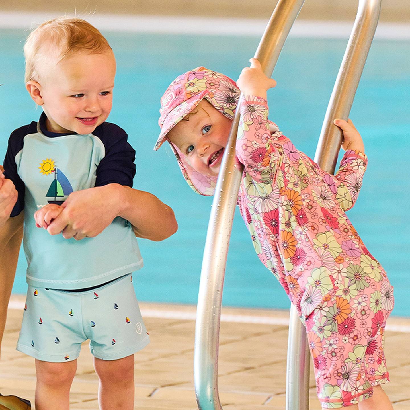 Zwei Kleinkinder lächeln an einem Pool; das eine in einem blauen Badeanzug hält den Arm eines Erwachsenen, das andere in einem geblümten Outfit klettert eine Leiter hinauf.