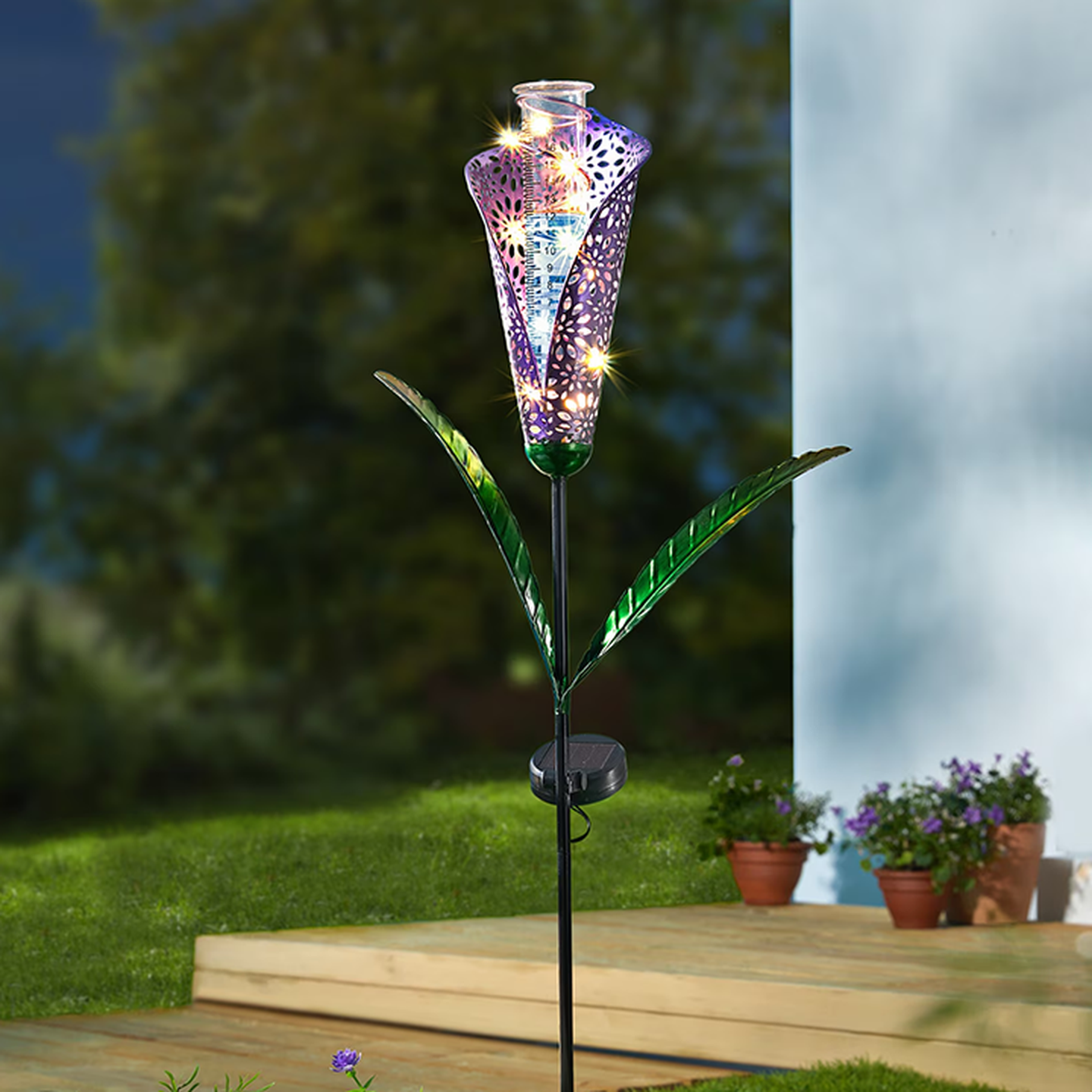 Eine solarbetriebene Gartenleuchte in Form einer bunten Blume mit leuchtenden Mustern, auf einem Rasen platziert, mit Topfpflanzen in der Nähe.