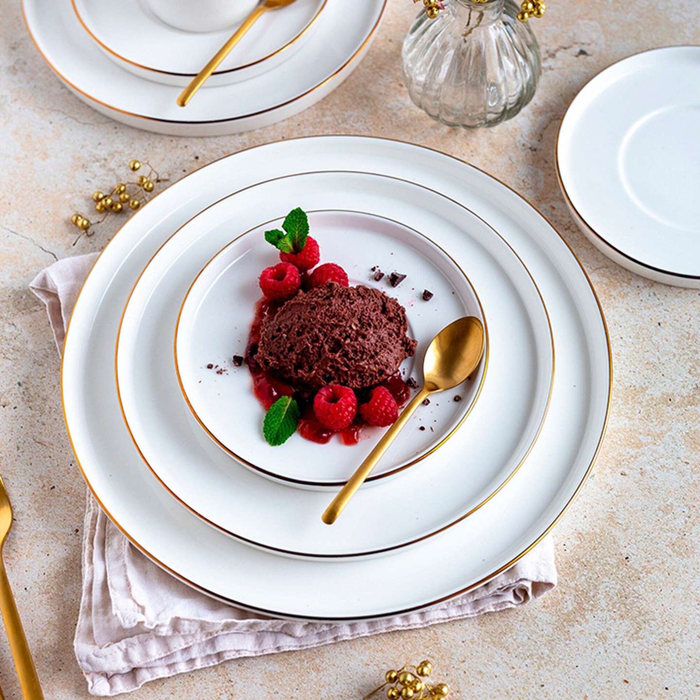 Ein elegantes Dessert-Arrangement mit Schokoladenmousse, Himbeeren und Minze auf feinem Porzellan, begleitet von einem goldenen Löffel, auf einer beigen Tischdecke.
