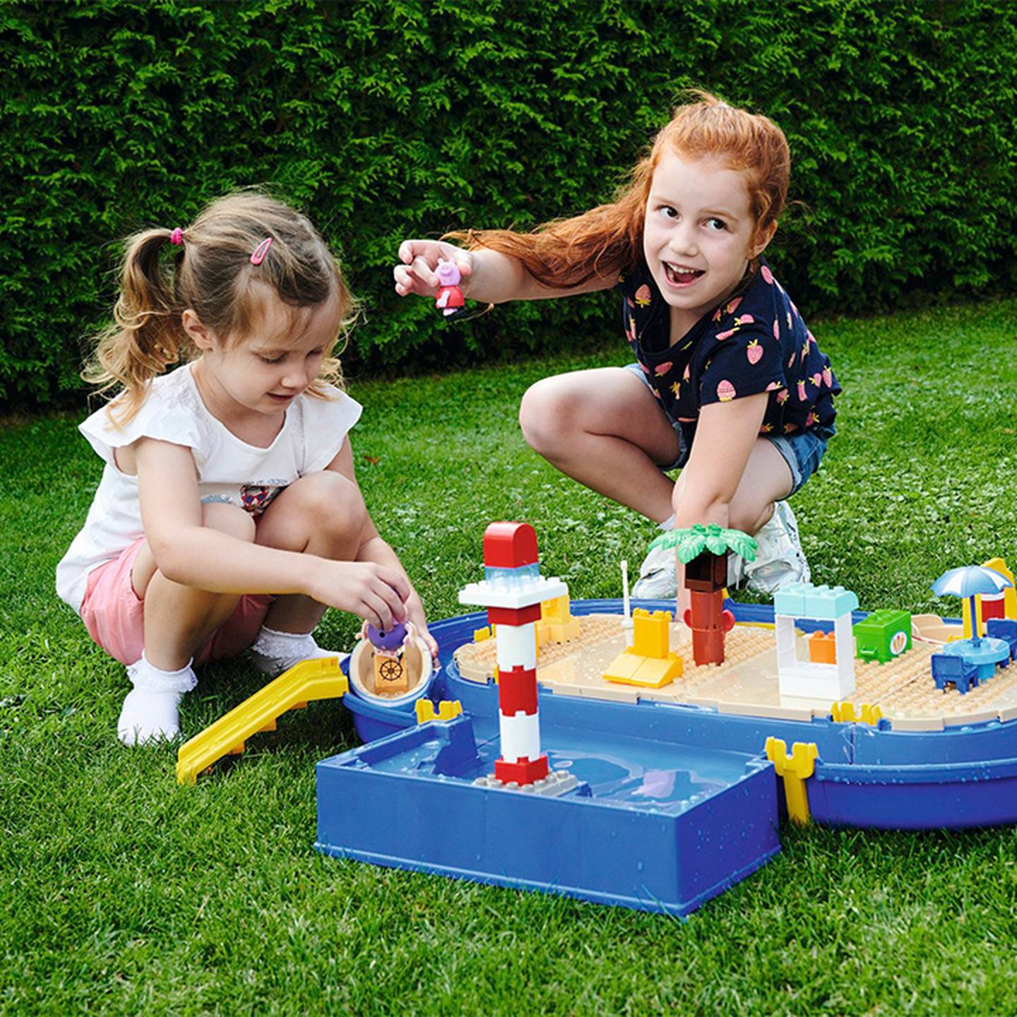 Zwei Kinder spielen auf der Wiese mit einem Spielzeug-Wasserpark, der einen Leuchtturm, eine Rutsche und Wasserspiele enthält.